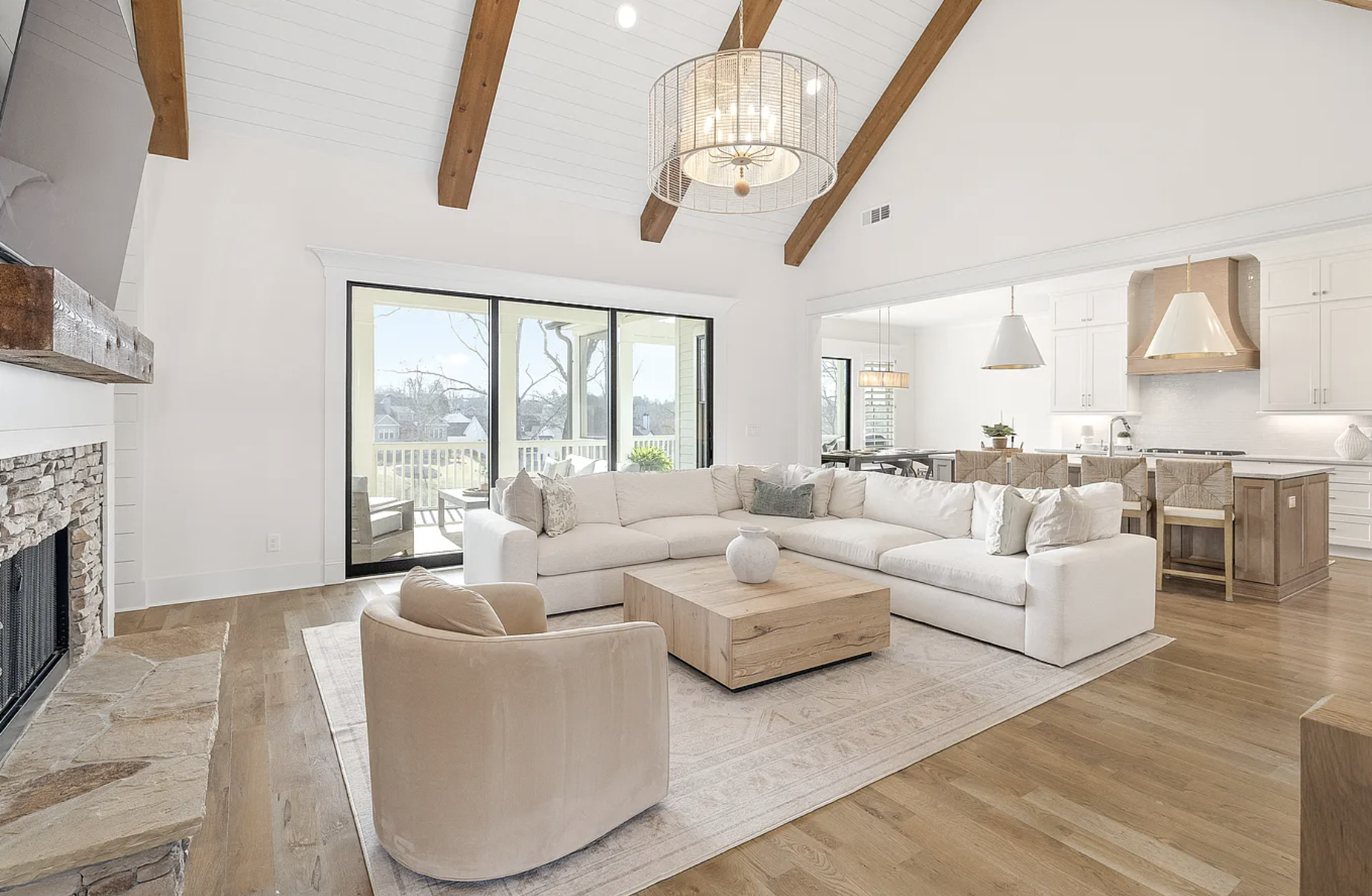 Bright living room with white L-shaped sofa, wooden coffee table, beige armchair, stone fireplace, large sliding glass doors leading to balcony, and open kitchen with white cabinets and pendant lighting.