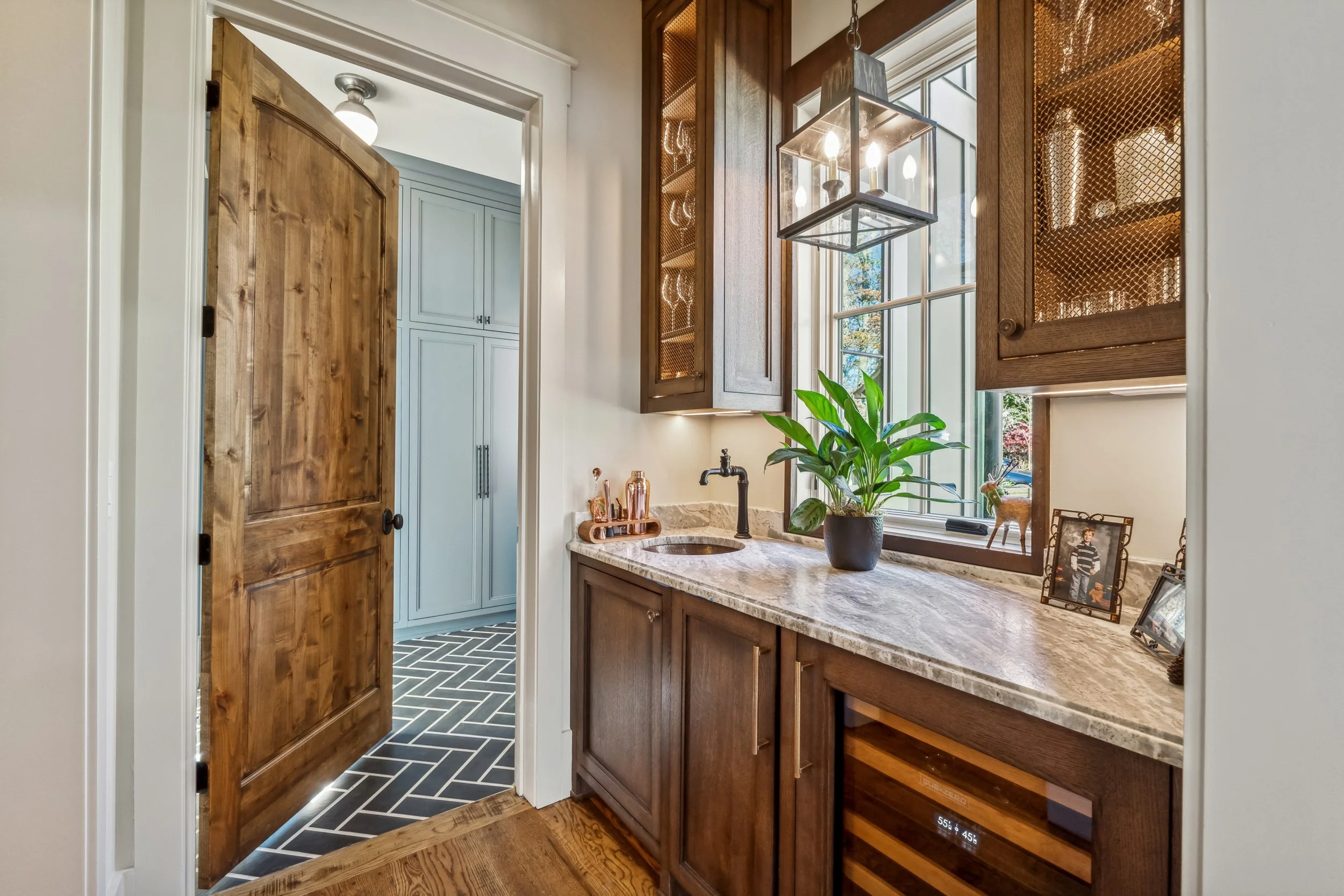 A kitchen with wooden cabinets, a granite countertop, a small sink, a potted plant, framed photos, and a window with blinds. There is a doorway leading to a laundry area with light blue cabinets and black and white herringbone patterned flooring.
