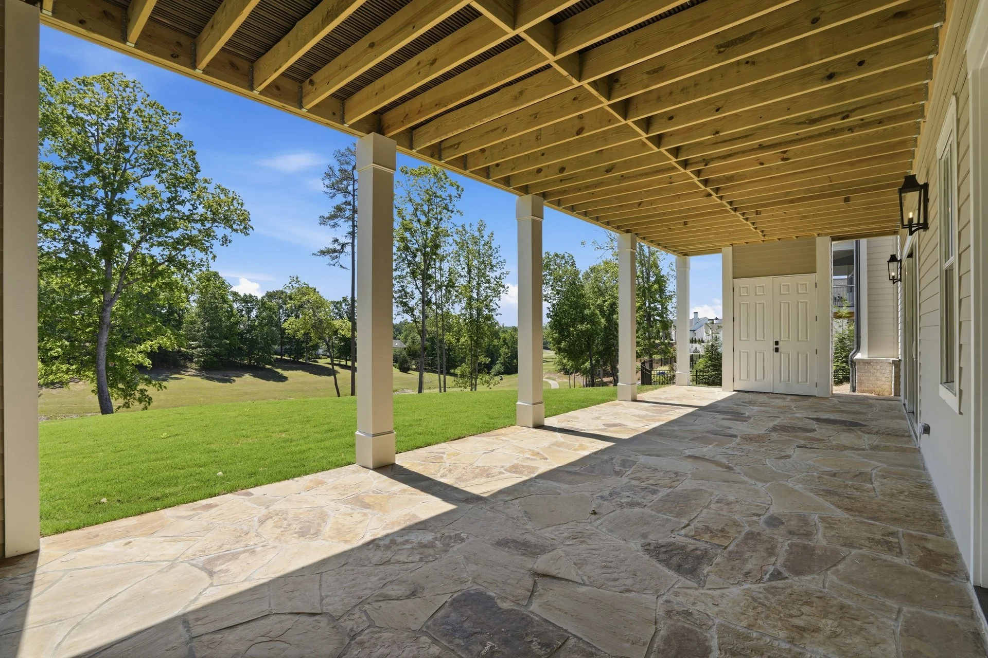 View from a covered porch with a stone floor, white columns, overlooking a grassy backyard with trees and a blue sky.
