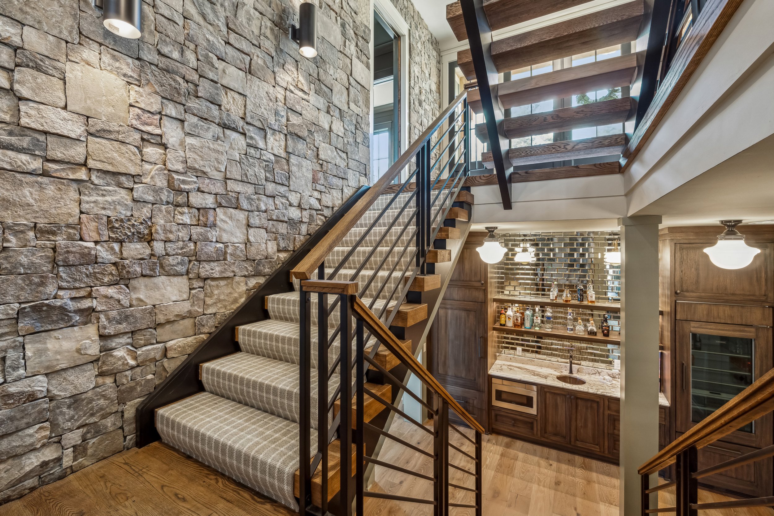 Interior view of a staircase with wooden steps and metal railing, next to a stone wall, leading to an upper floor with large windows, and a bar area with bottles and glasses in the background.