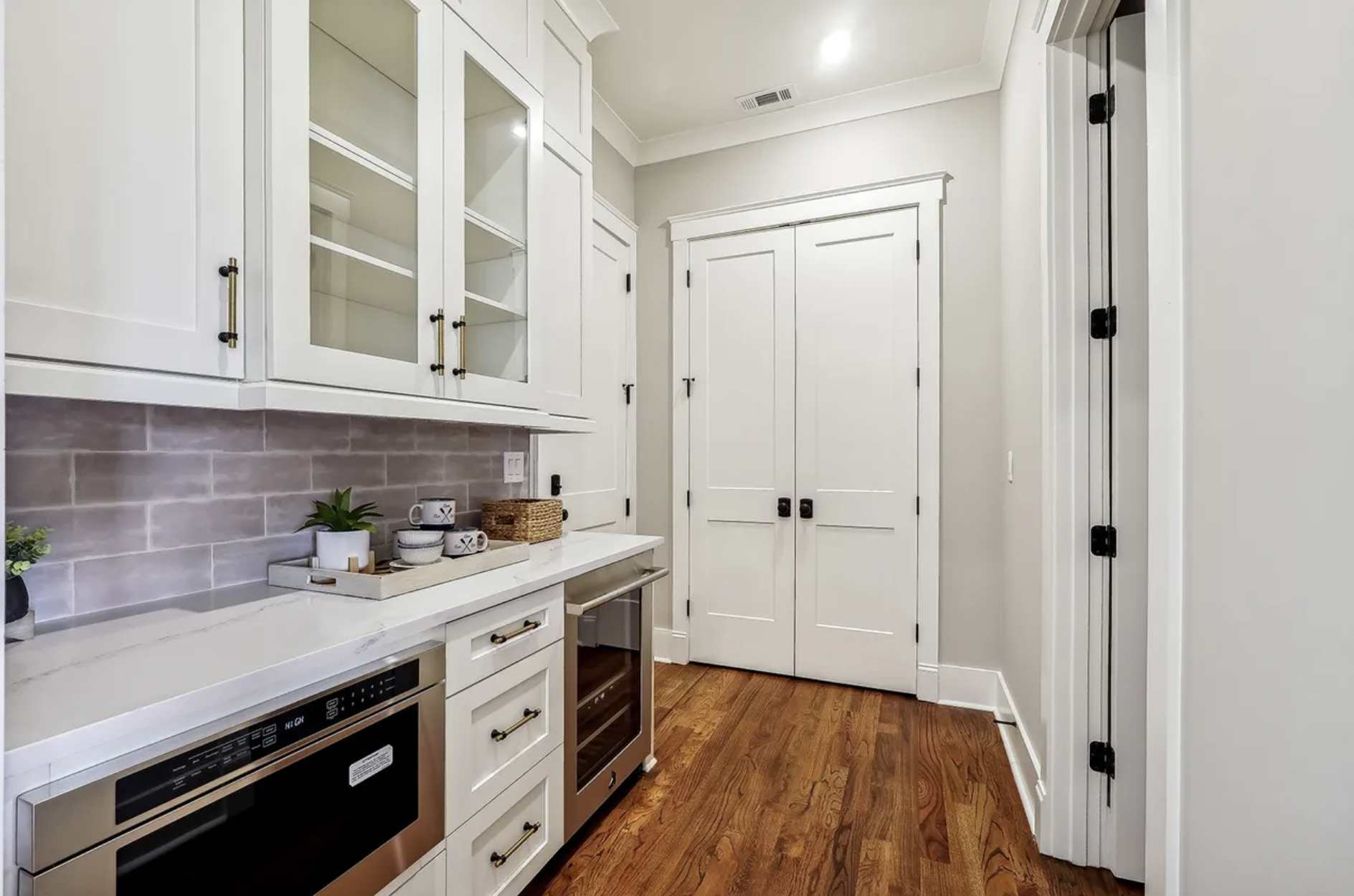 A modern kitchen with white cabinets, glass-front upper cabinets, a granite countertop, and a white subway tile backsplash. There are small plants and mugs on the counter. The floor is wooden, and there are double doors and a laundry or utility door.