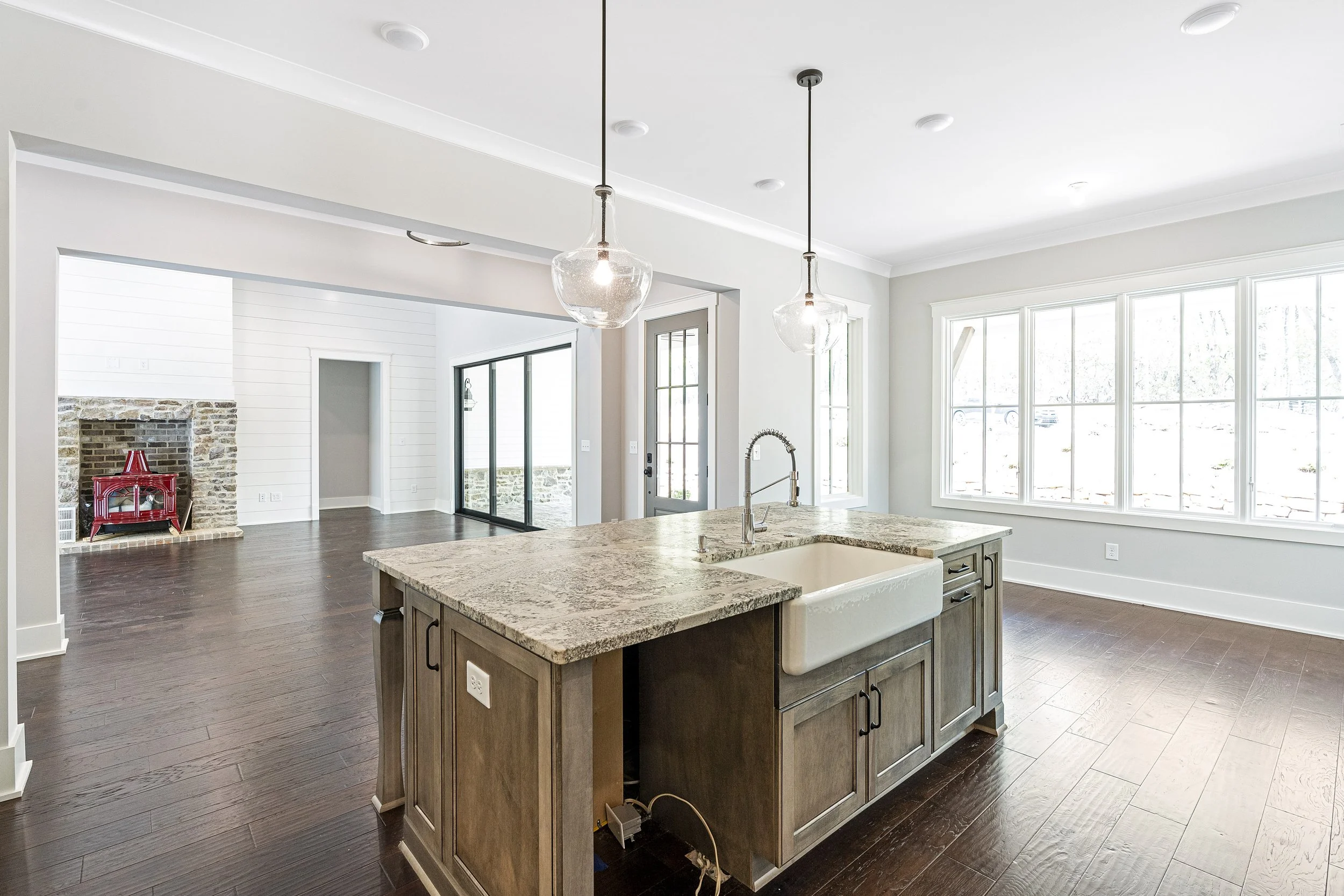 A modern kitchen with a large island with a granite countertop, a farmhouse sink, and dark wooden cabinets, overlooking a bright living area with large windows and a sliding glass door leading outside.