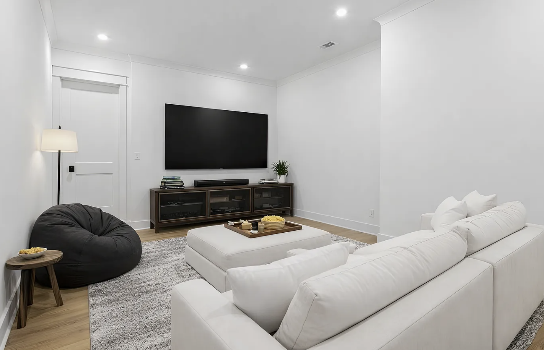Modern living room with white sectional sofa, black bean bag chair, wood TV console, mounted flat-screen TV, and minimal decor.