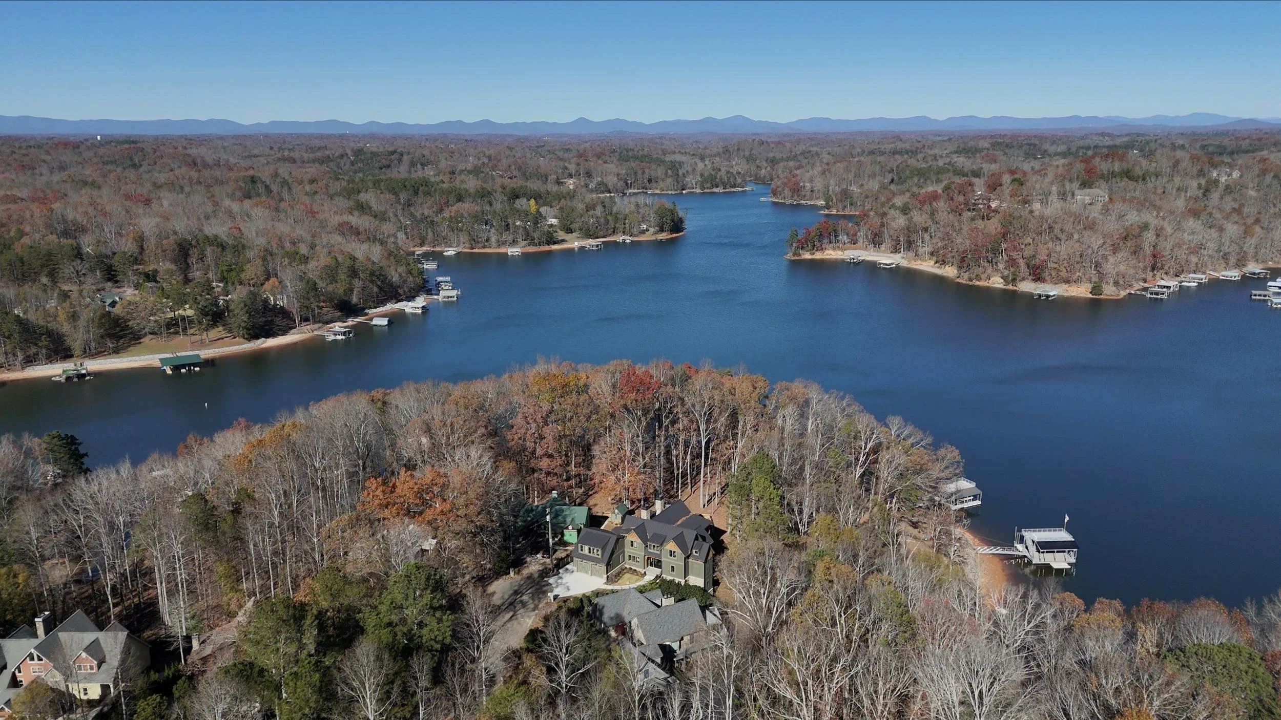 Aerial view of a lake surrounded by houses and trees with fall foliage, mountains in the distance under a clear blue sky.