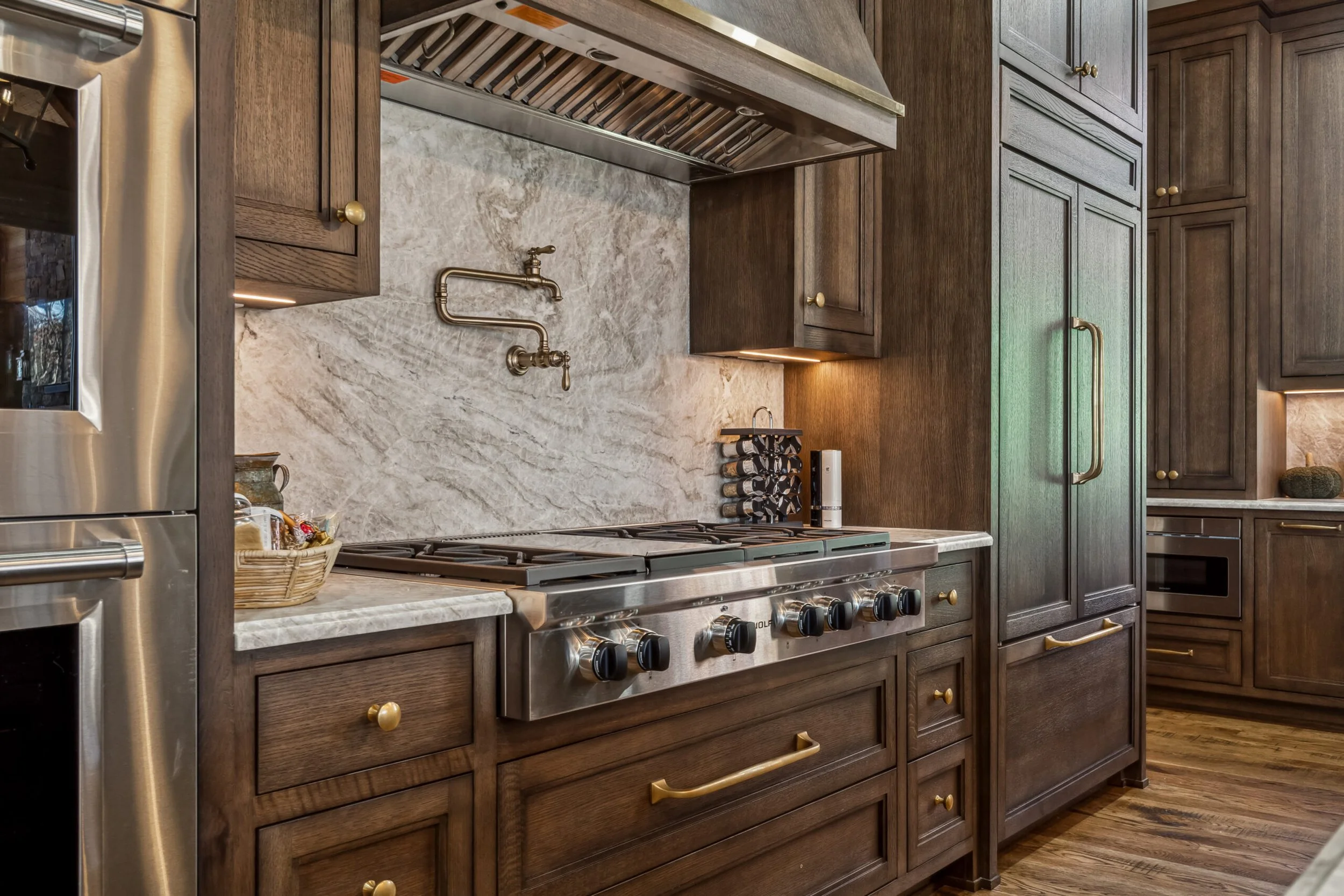 Kitchen with dark wooden cabinets, marble countertops, and a stainless steel stove with black knobs. There is a basket of spices and a wine rack with bottles on the counter. A brass faucet is above the stove on a marble backsplash. There is a vent ho