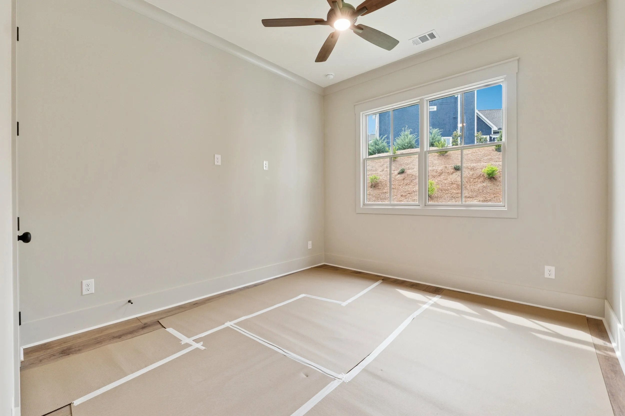 Empty room with beige walls, large window, ceiling fan with lights, and taped outline on the carpet for flooring work.