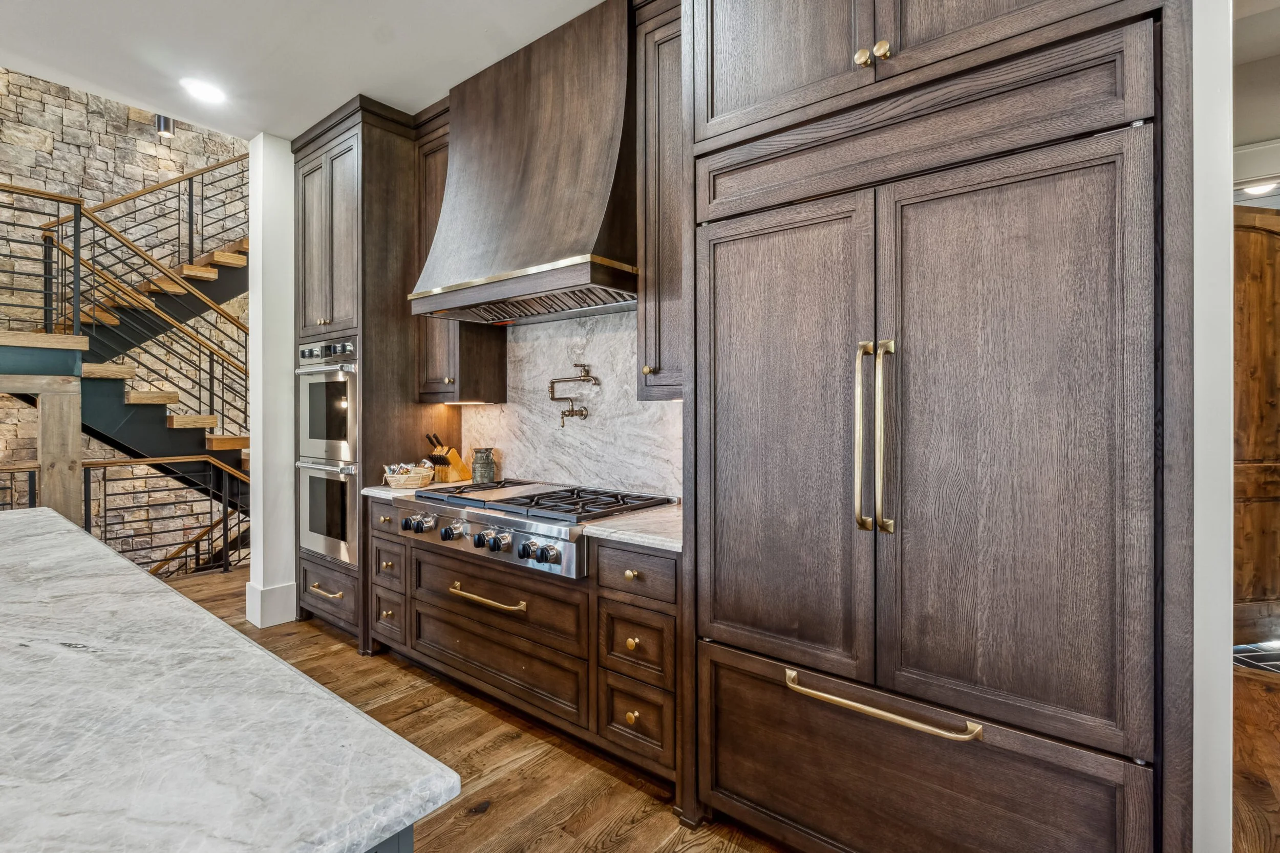 Modern kitchen with dark wood cabinets, a stainless steel stove, oven, and a large vent hood. Stone accent wall with stairs and black railings are visible in the background.