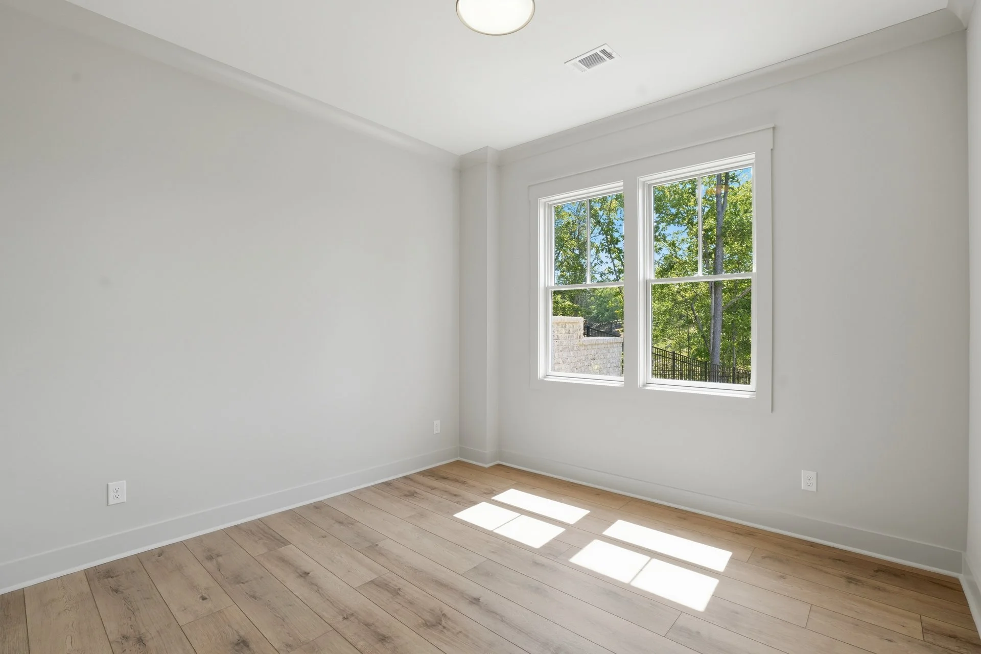 Empty room with light-colored hardwood floors, white walls, and large windows showing trees outside.
