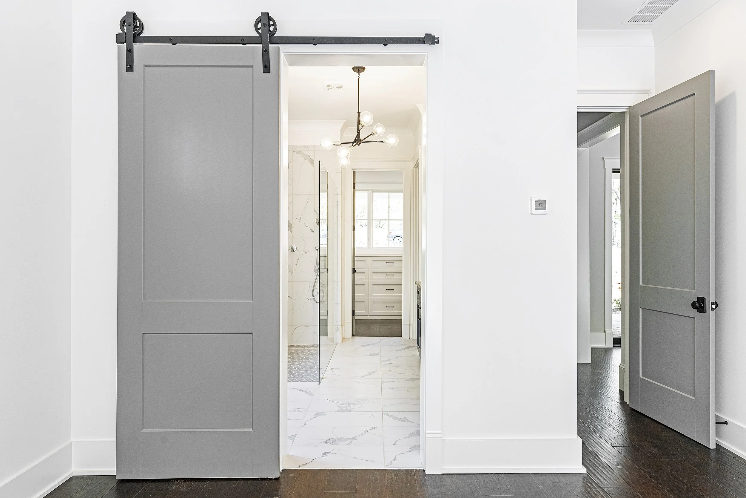 A view of a modern home interior with a kitchen and a bathroom visible through doorways. The space features white walls, dark hardwood floors, a gray sliding barn door, and a contemporary chandelier.