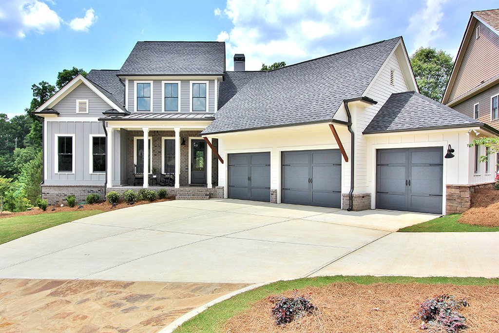 Modern house with gray and white exterior, three-car garage, front porch with chairs, landscaped yard, and blue sky.