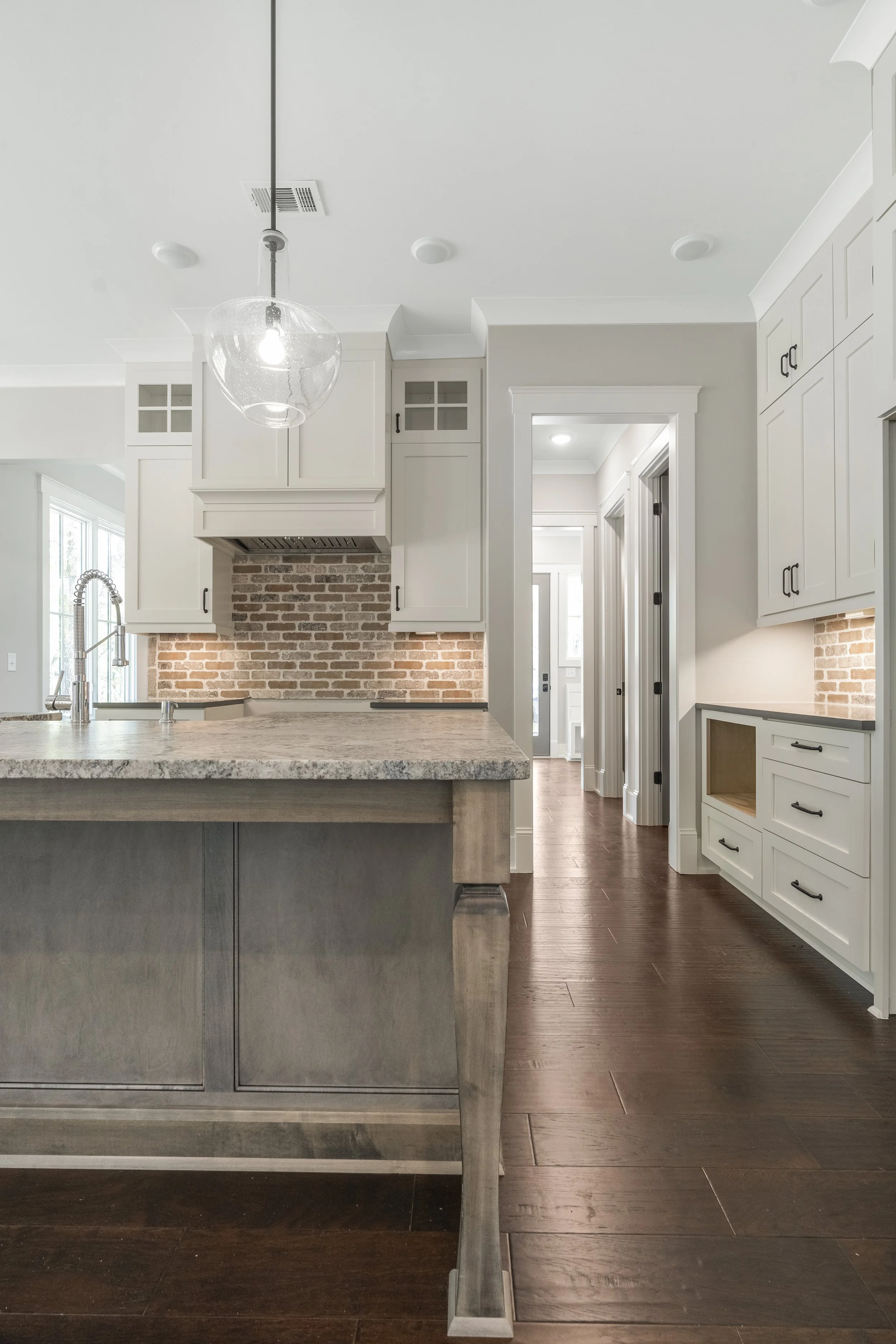 A kitchen with white cabinets, brick backsplash, wooden floor, and a large kitchen island with a marble countertop. A glass pendant light hangs from the ceiling.