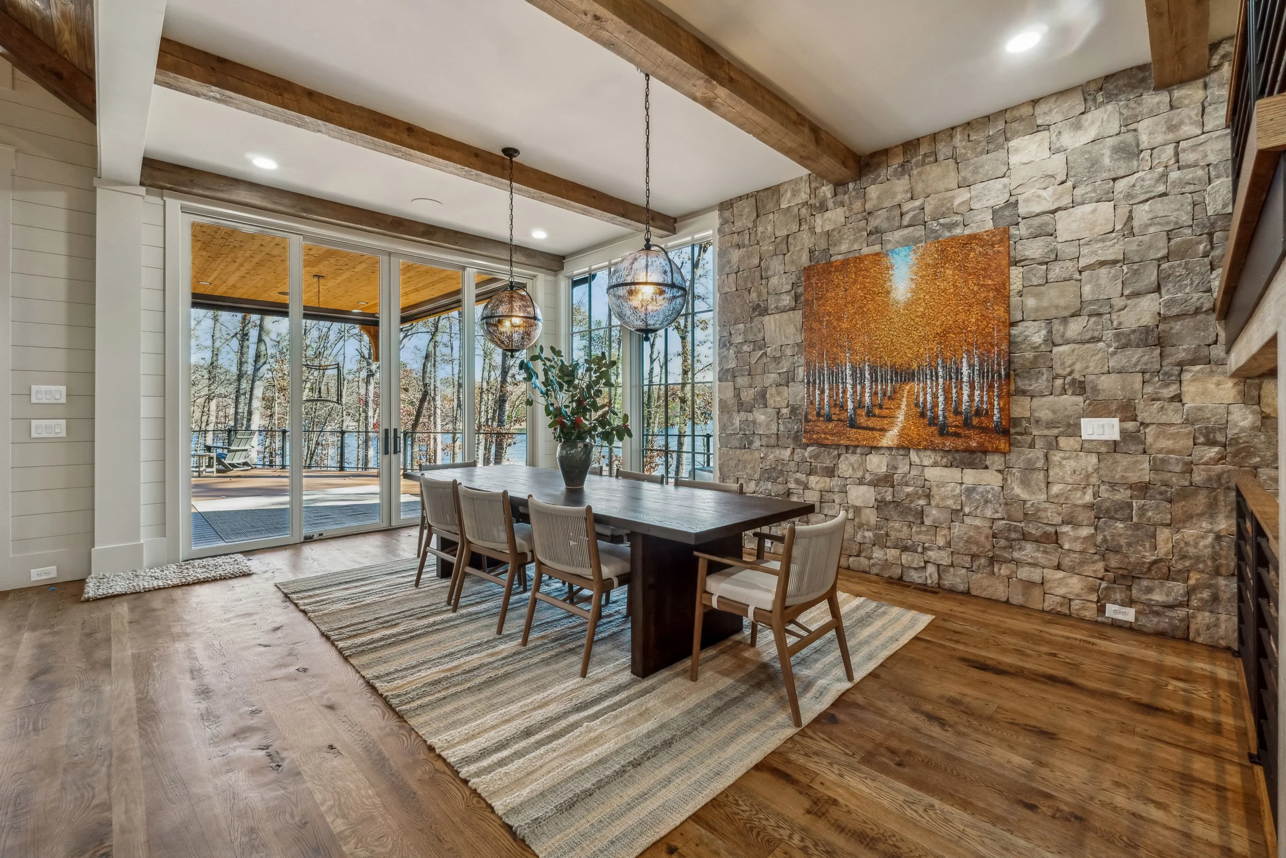 A cozy dining room with a wooden table, eight chairs, hardwood flooring, a stone accent wall with a colorful painting, two hanging pendant lights, a rug, large sliding glass doors leading to an outdoor deck, and a window showing trees outside.