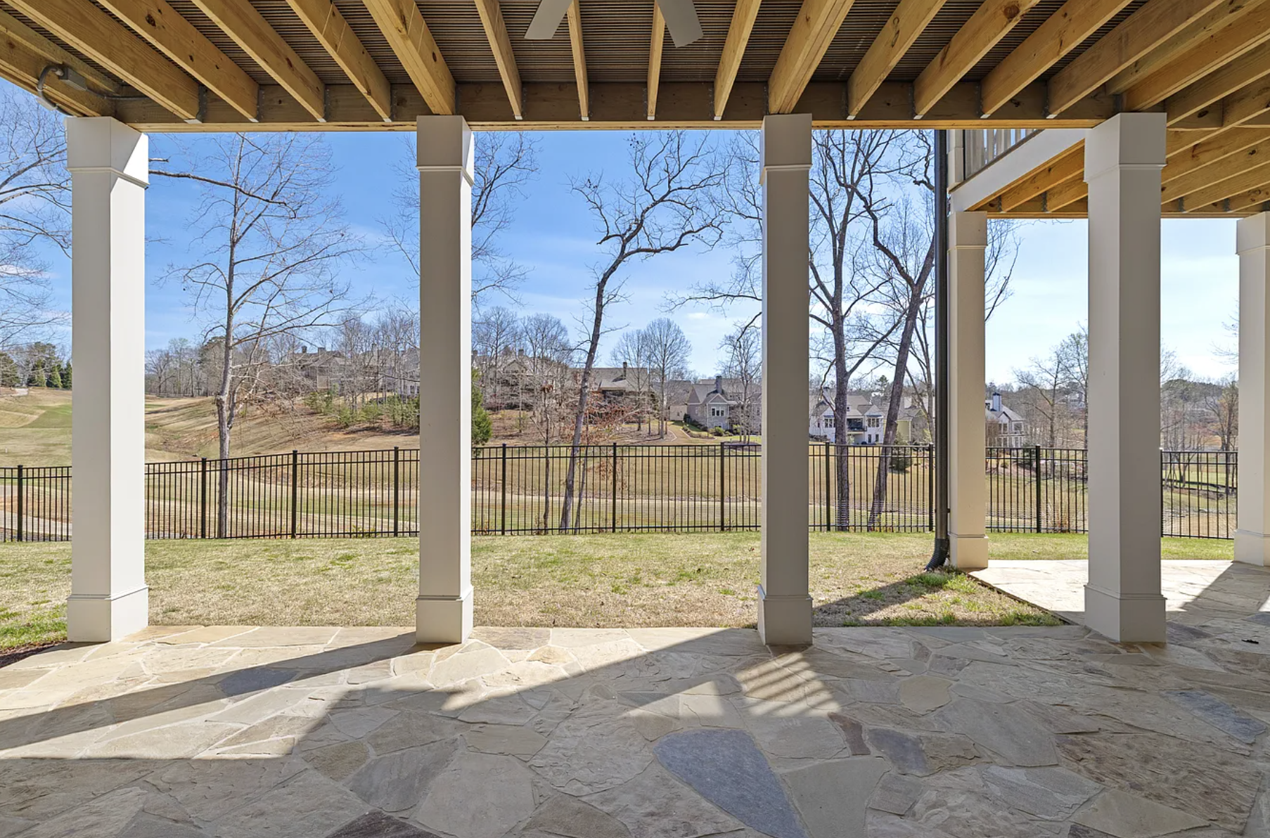 View through a covered porch showing a backyard with a black metal fence, trees, grass, and neighboring houses in the distance under a blue sky.