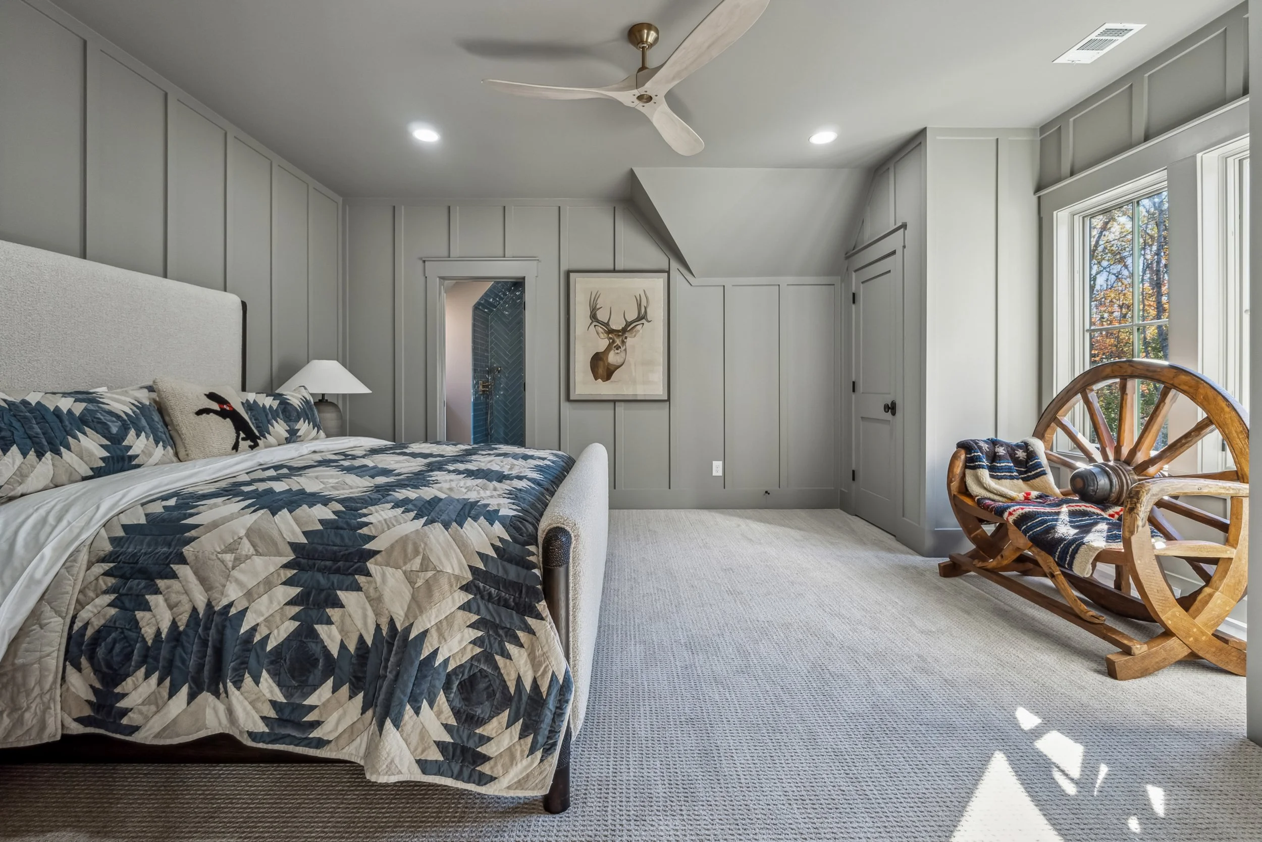 Bedroom with a bed, quilt, and pillows, a nightstand with a lamp, a deer portrait on the wall, a window showing fall foliage, a rocking chair with a blanket, and a ceiling fan.