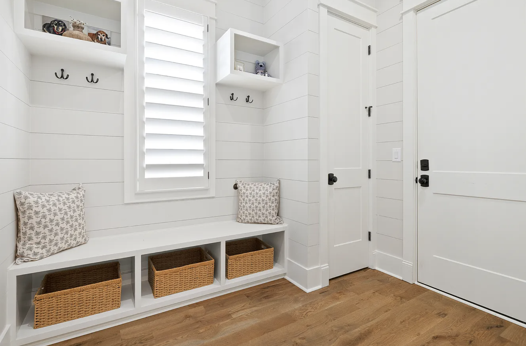 A white mudroom with a window that has white shutters, built-in white benches with two decorative pillows, and three woven baskets in cubbies underneath the bench. There are black hooks on the wall, and two white open shelves with decorative items. T