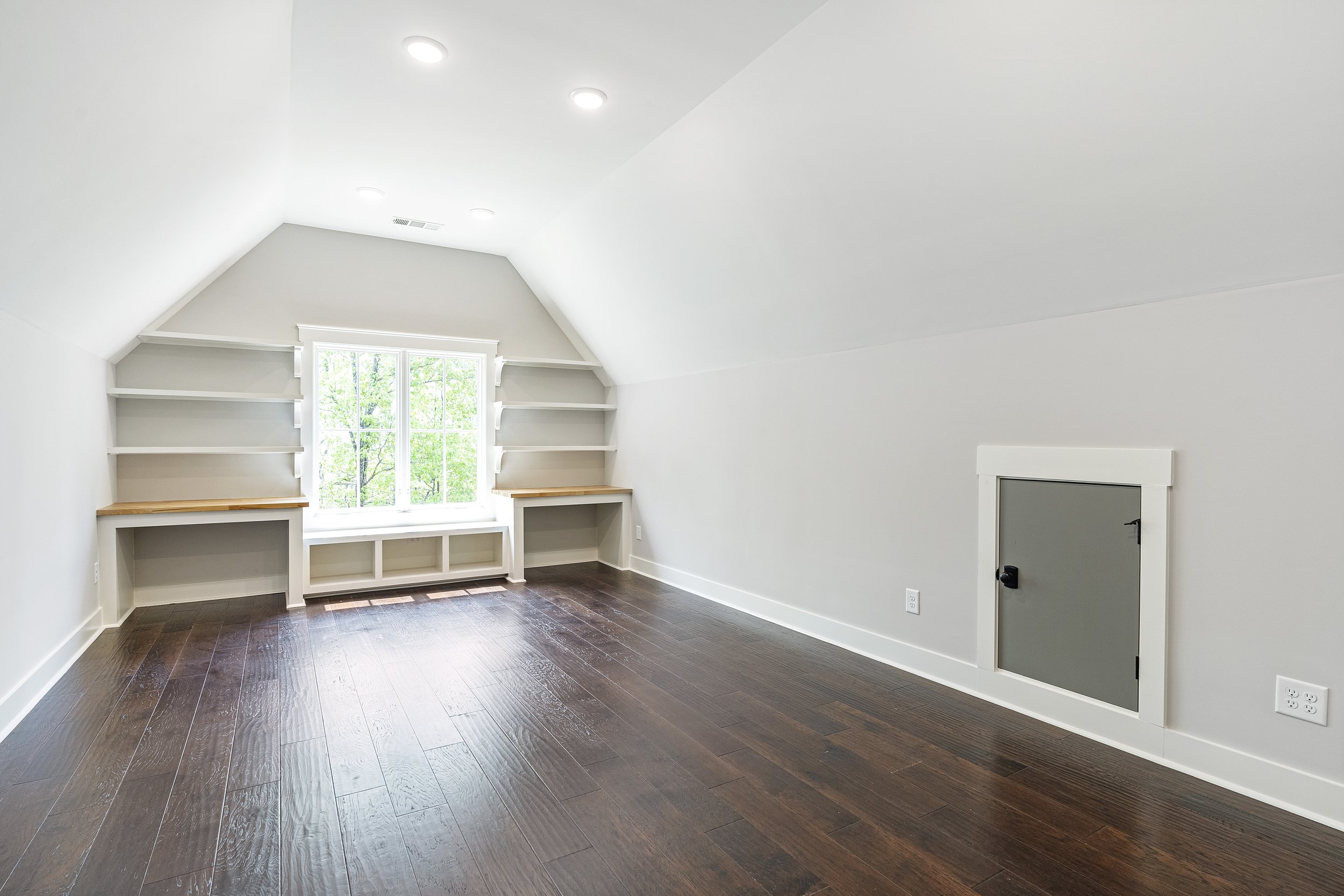 An empty room with white walls, dark wood flooring, a window with trees outside, built-in shelves under the window, and a small door in the wall.