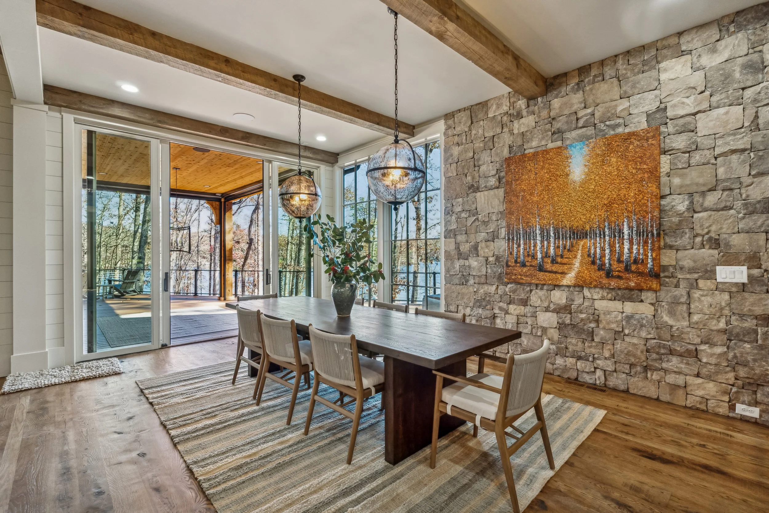 A dining room with a wooden table surrounded by six matching chairs on a striped beige and blue rug. There is a large abstract painting of a forest on a stone wall. Two hanging globe pendant lights with rustic metal finishes are suspended above the t