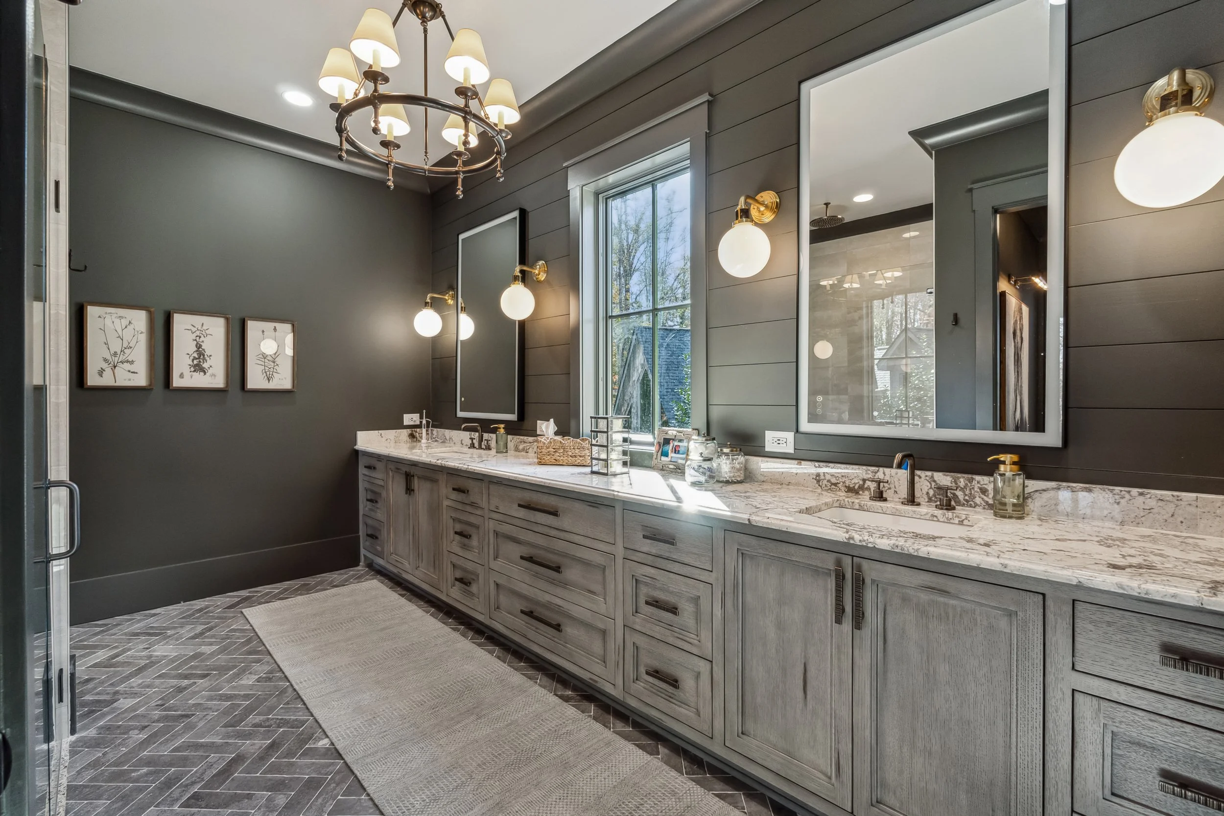 Modern bathroom with gray walls, wooden vanity with a white marble countertop, large mirrors, and contemporary lighting fixtures. Art prints are on the wall, and a window lets in natural light.