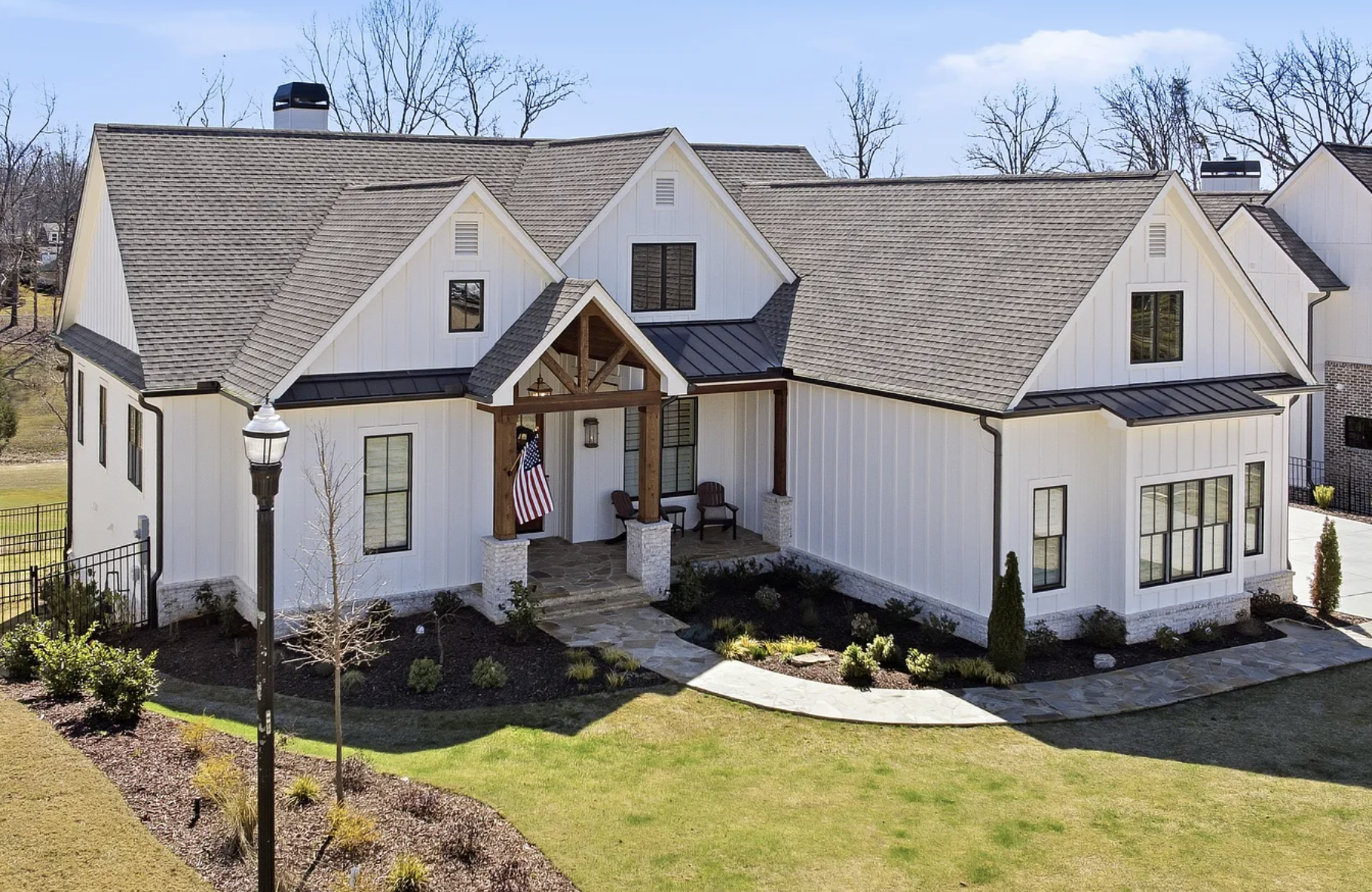 A modern white house with a gabled roof, black windows, and a small porch with wooden beams and stone supports, surrounded by a landscaped yard with trees, shrubs, and a stone walkway.