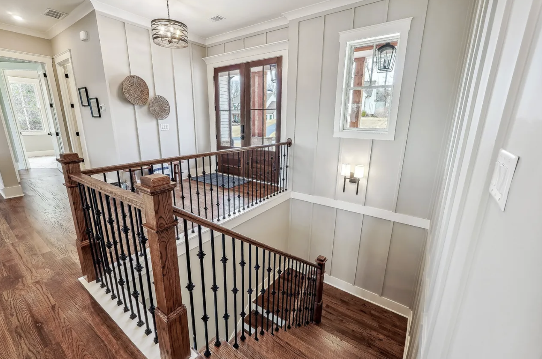 Interior view of a house landing with wooden flooring and white paneled walls. There is a wooden front door with glass inserts, flanked by a window on the right. A wall-mounted lamp is next to the window. Woven decorative baskets hang on the wall, an