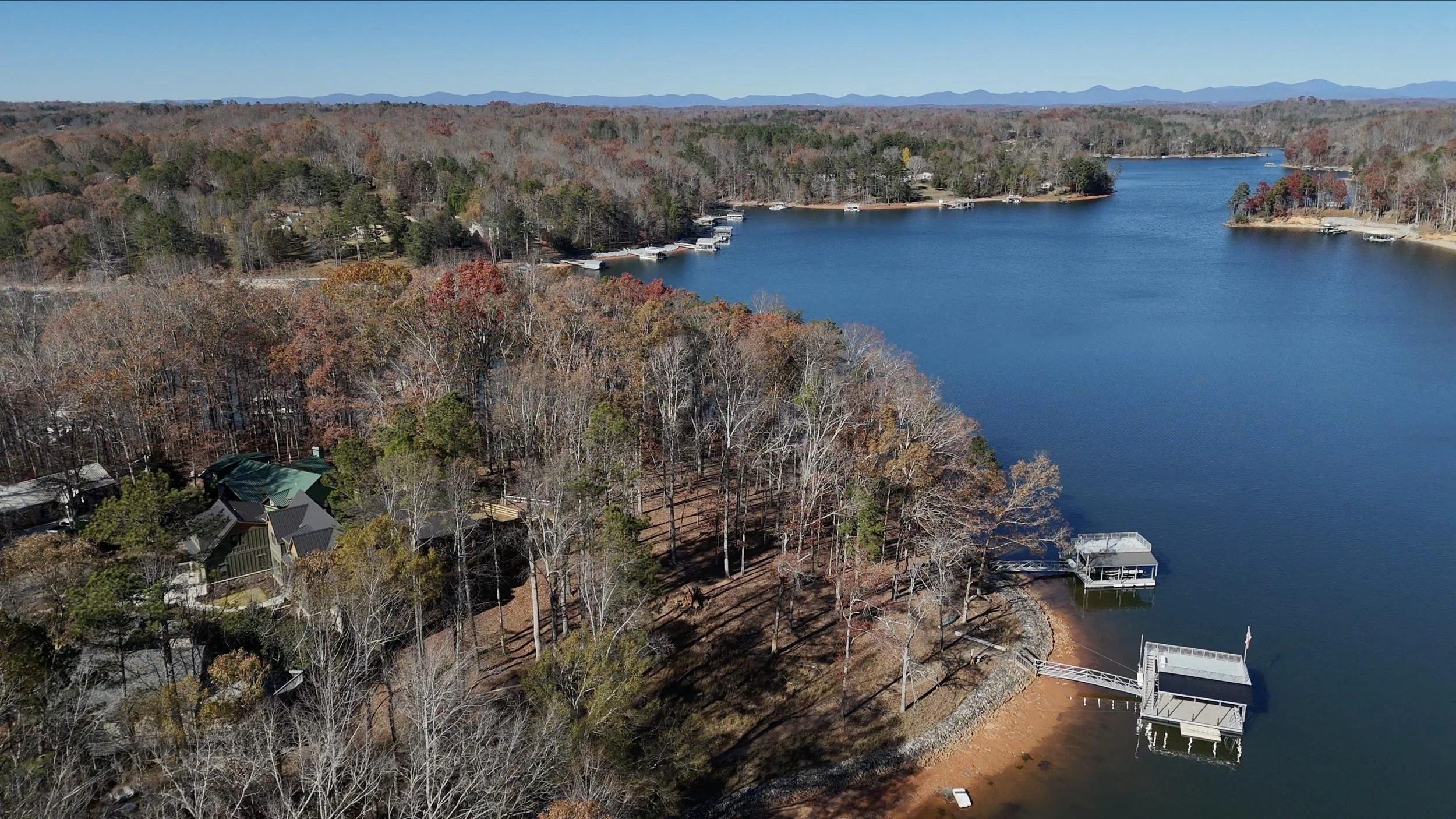 Aerial view of a lake with houses and docks along the shoreline, surrounded by forest with trees in autumn colors, under a clear blue sky.