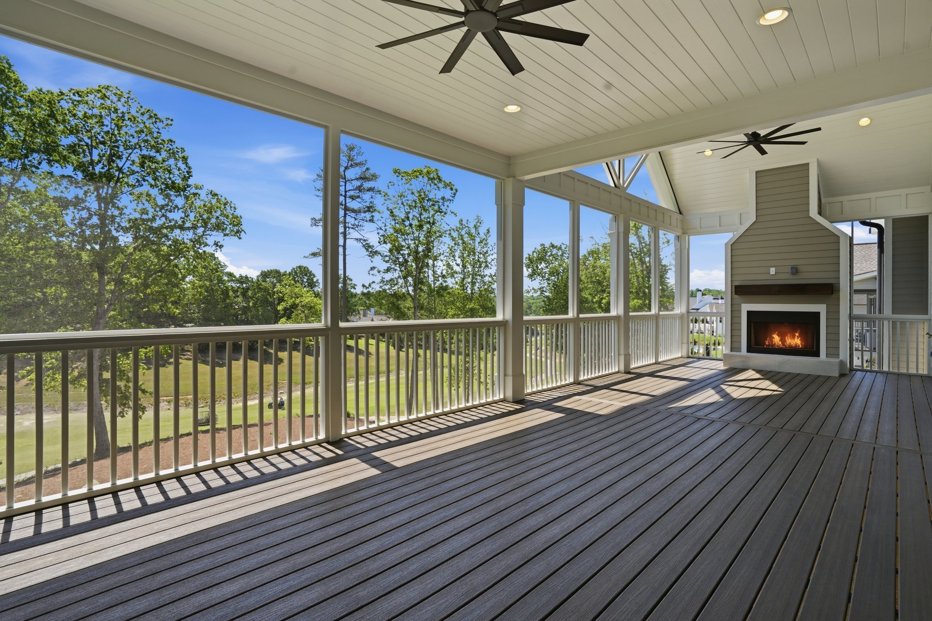 Covered balcony with wooden flooring, white railing, ceiling fans, and a fireplace, overlooking green trees and a blue sky with clouds.