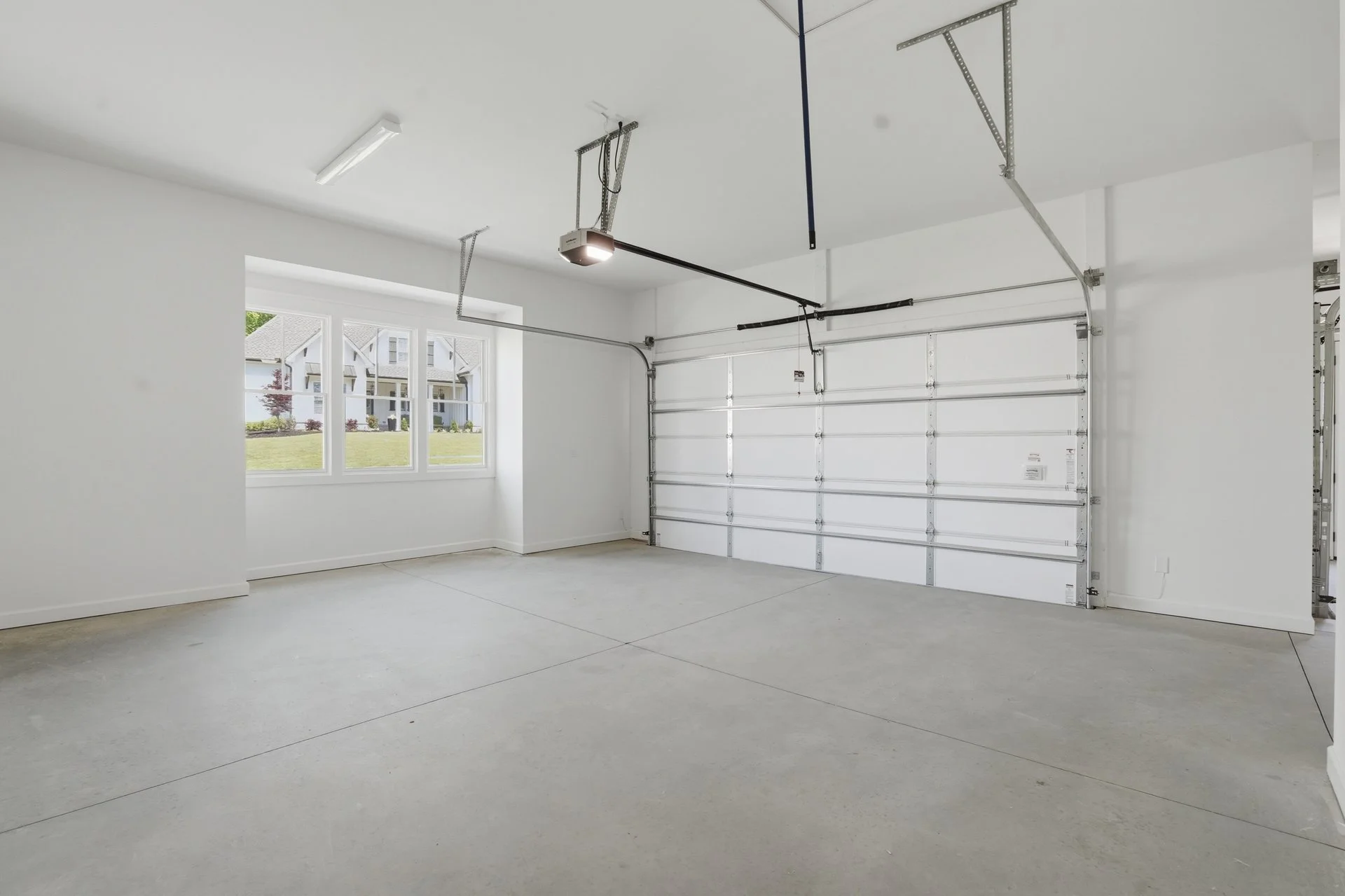 Empty white garage with a large window showing a suburban neighborhood outside, a closed white garage door, and concrete floor.