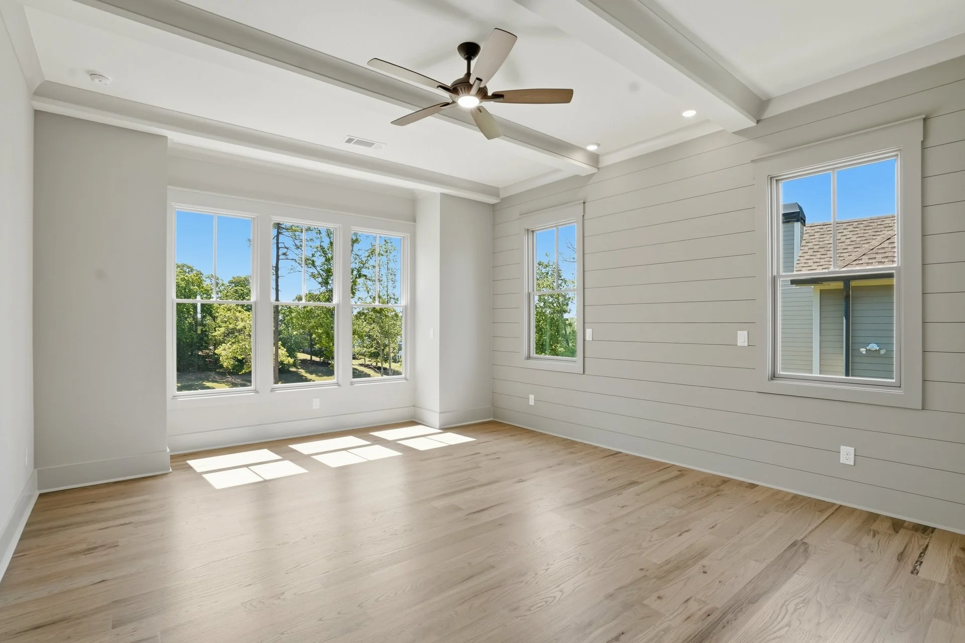 Empty modern room with large windows, white walls, ceiling fan, and light wood flooring.