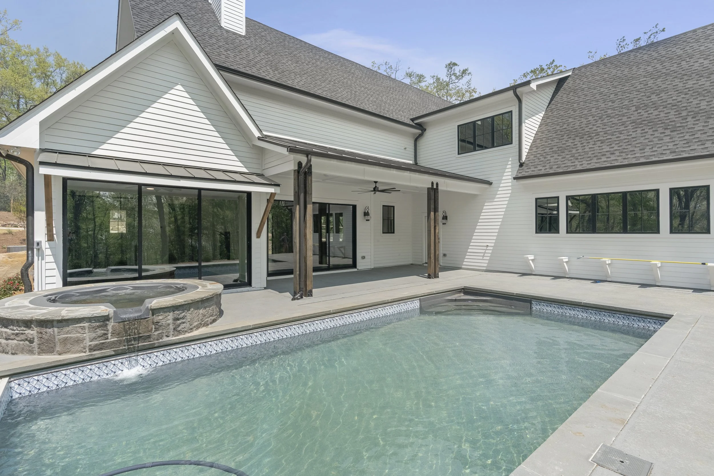 A modern house with a backyard pool, hot tub, and a covered patio area. The house features white siding, large black-framed windows, and a sloped roof. The scene is set on a clear day with trees in the background.