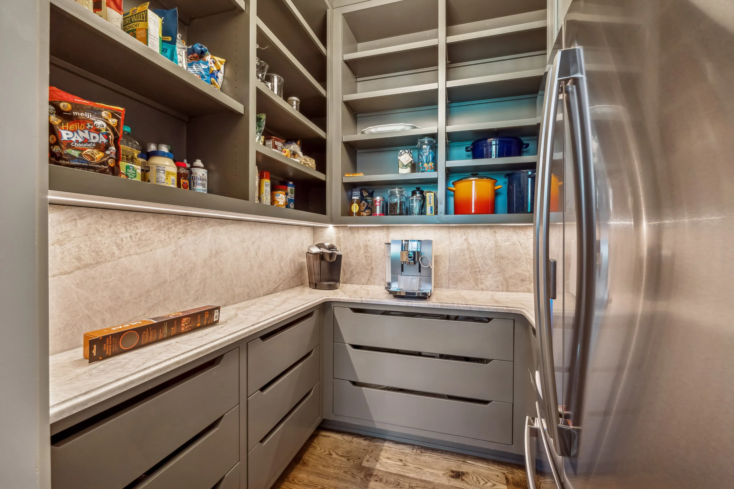 A walk-in pantry with gray cabinetry and open shelves filled with snacks, jars, and kitchen items. A marble countertop runs along the bottom with a coffee machine and a box. The walls and shelves are gray, and the floor is wood. Part of a stainless s