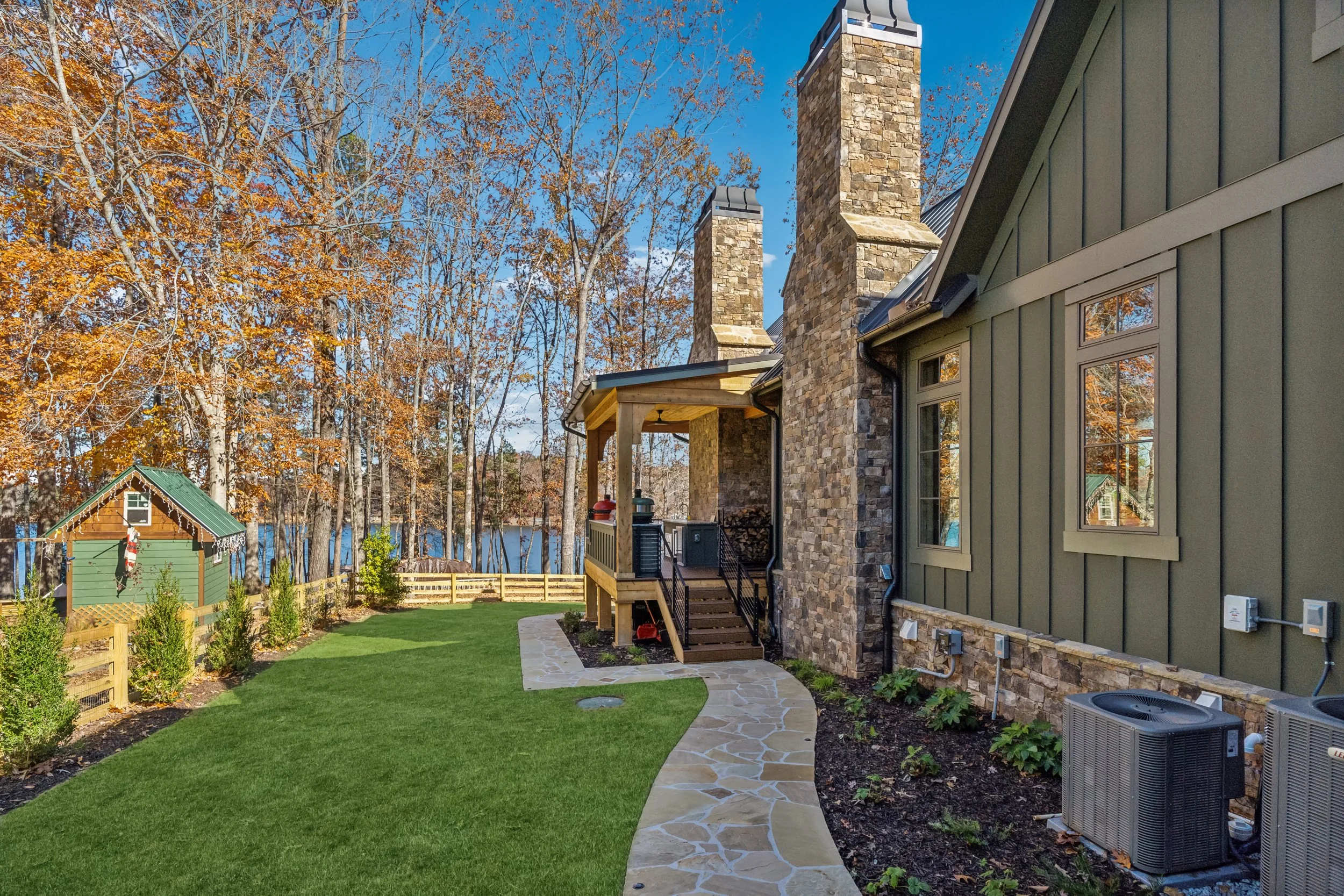 Backyard with well-maintained green grass, stone pathway, house with green siding and stone chimney, trees with autumn leaves, and a small playhouse with holiday decorations.