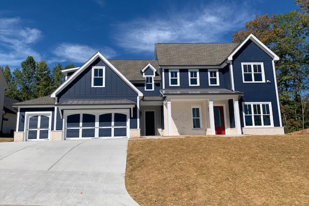 A modern two-story house with a blue exterior, multiple windows, a covered front porch with white columns, and a garage with white doors. The house is situated on a sloped lawn with a concrete driveway, under a partly cloudy sky.