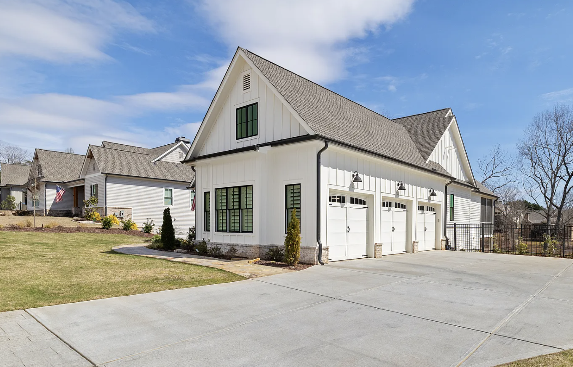 A white two-story house with a front garage, black window frames, and a gray shingle roof, surrounded by a lawn and neighboring houses with American flags.