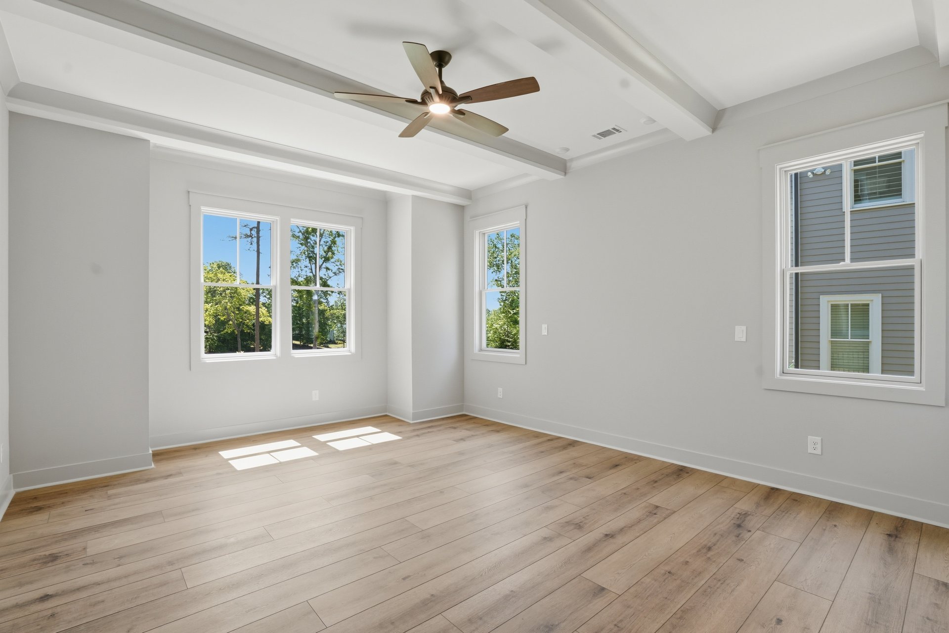 Empty whiteroom with large windows, light wood floors, ceiling fan, and natural sunlight filtering in.