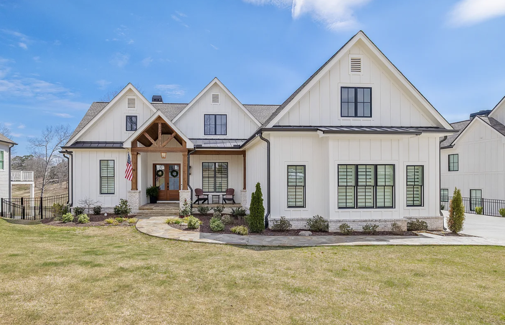 A modern white house with a dark roof, front porch with wooden accents, and a landscaped yard with a stone walkway, small shrubs, and lawn.