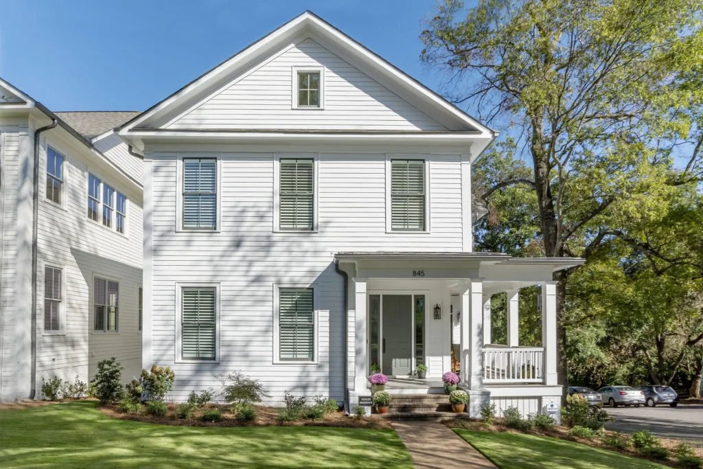 A white two-story house with a front porch, potted plants with flowers, and a well-maintained lawn, with trees and parked cars in the background.
