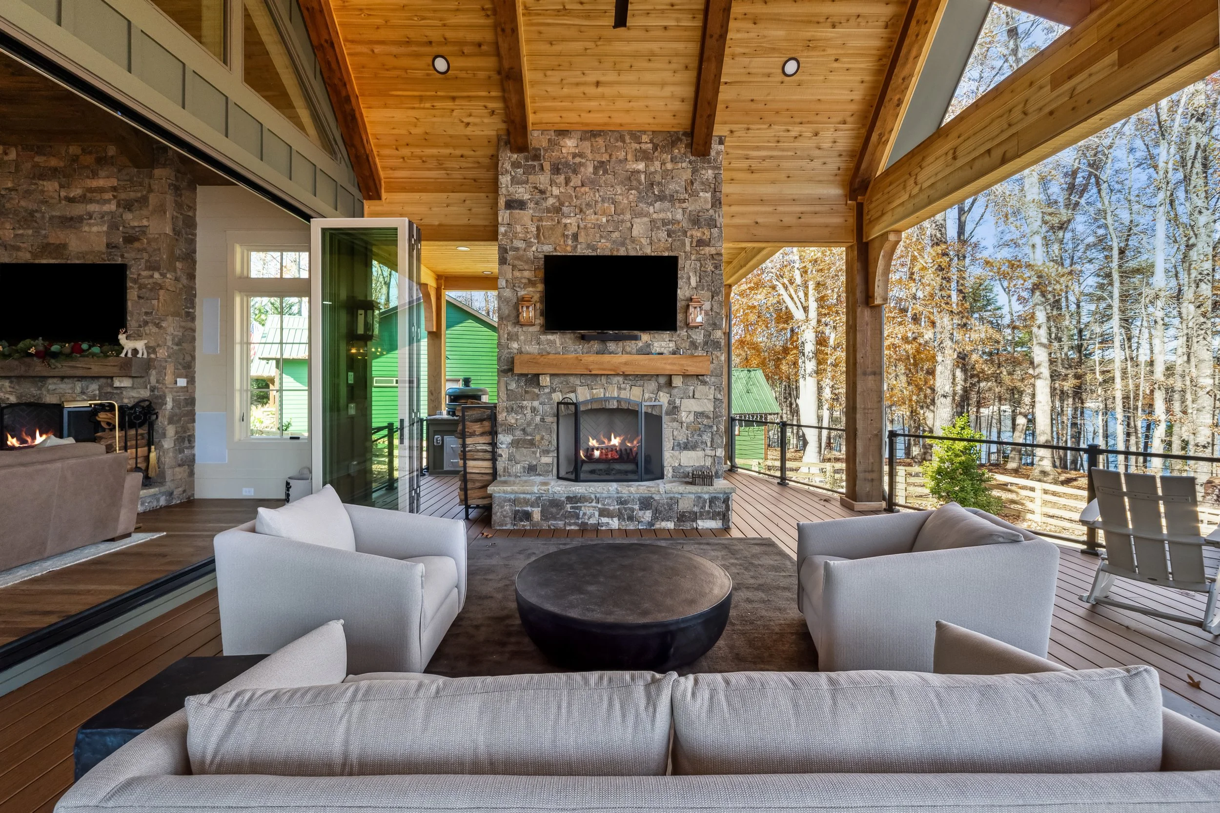 A spacious living area with a vaulted wooden ceiling, stone fireplace, and modern decor, opening to a deck with outdoor views of trees and neighboring houses.
