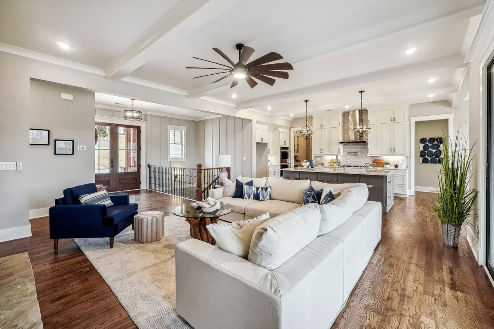 Open-concept living room with white sofa, navy armchair, wooden coffee table, and decor pillows. The space features hardwood floors, a kitchen with white cabinets, an island, and a brick chimney. There's a ceiling fan, recessed lighting, and large wi