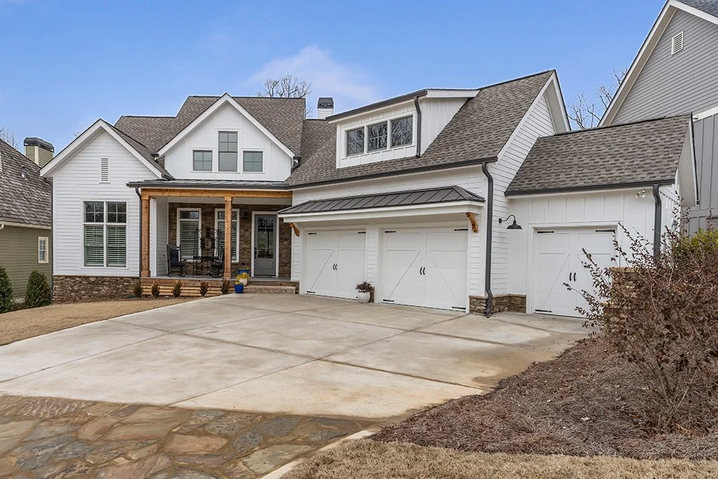 Front view of a two-story house with a white exterior, stone accents, and a three-car garage. There is a small covered porch with outdoor seating, and a paved driveway leading up to the garage.