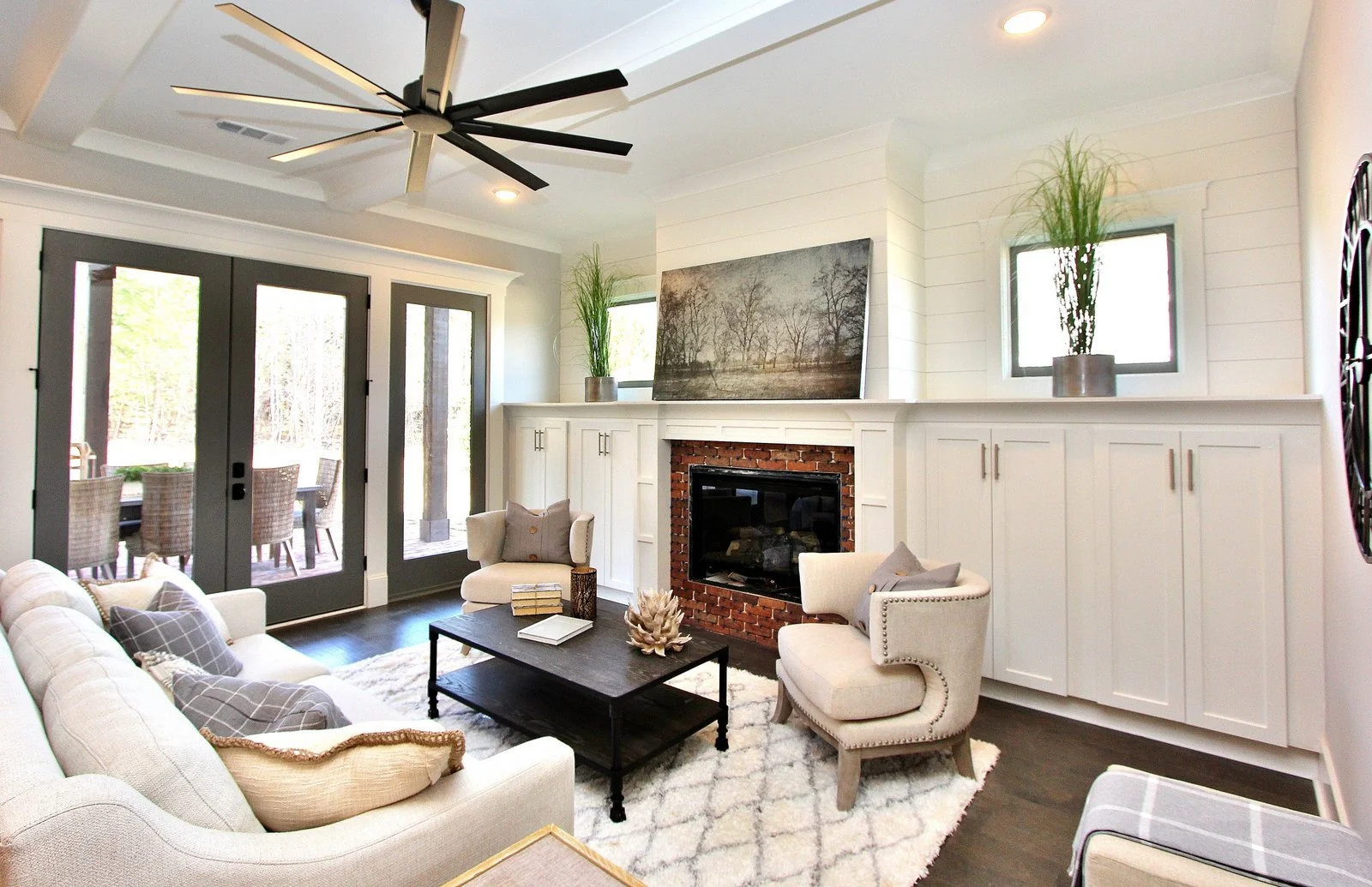 Living room with white walls, a fireplace with a brick surround, a large framed landscape painting above, two tall potted plants on either side of the painting, a beige armchair, a beige sofa with pillows, a dark coffee table with books and decorativ