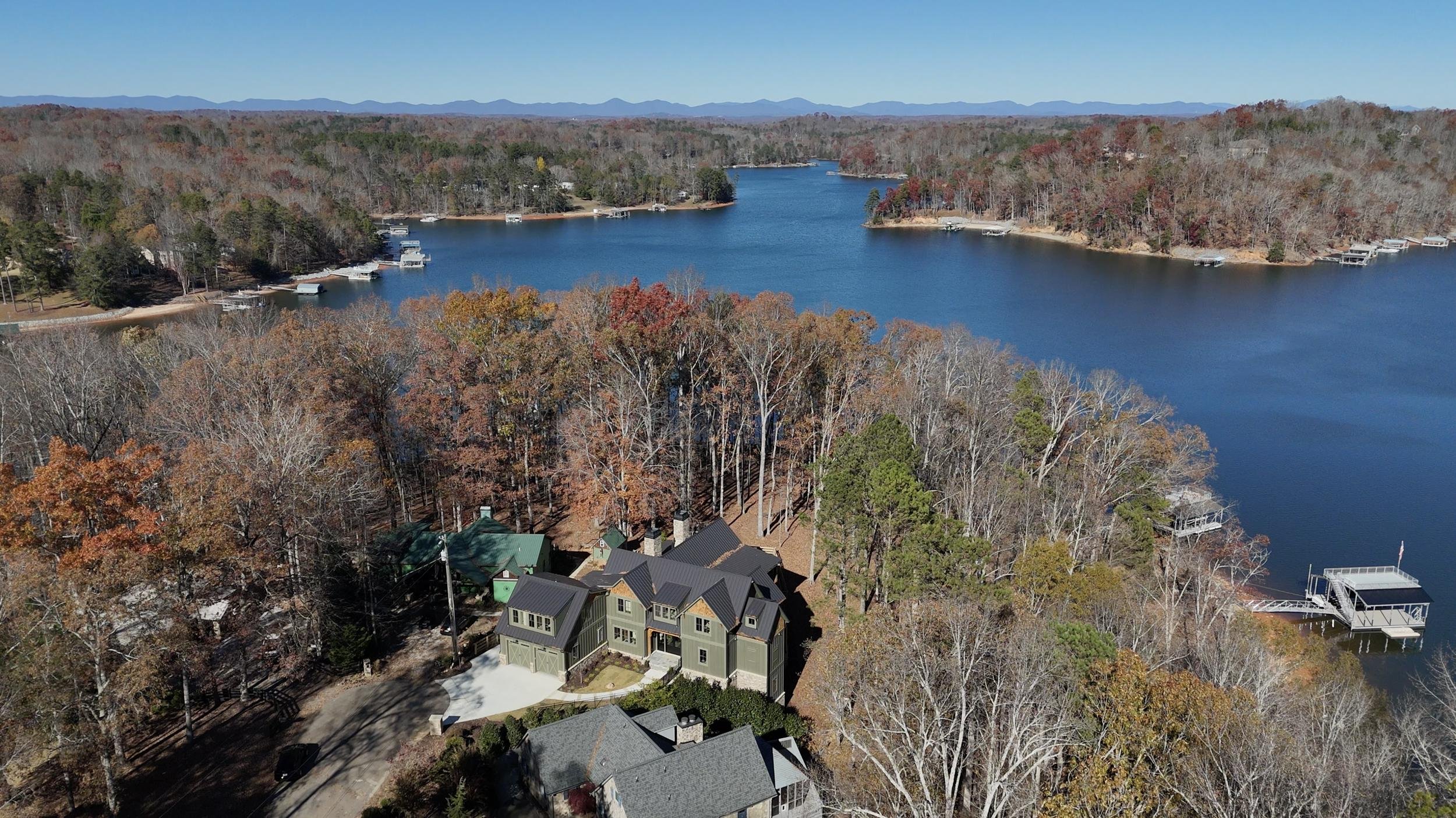 Aerial view of a lake surrounded by trees with fall foliage, houses and boats along the shoreline, and mountains in the distance under a clear blue sky.
