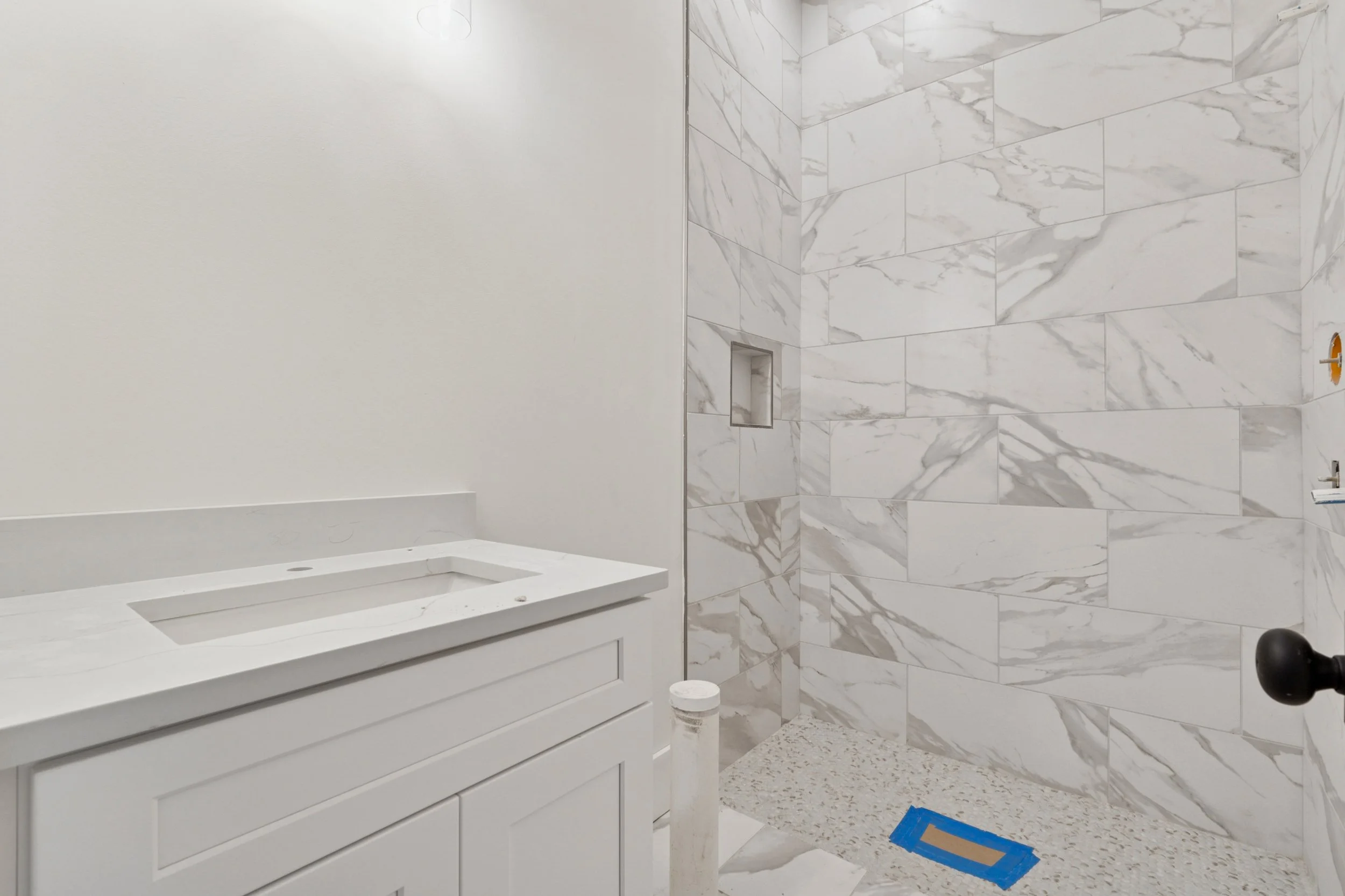 A bathroom with a white vanity sink, marble tile walls, and an empty shower area with a niche. The bathroom appears to be under construction or renovation.
