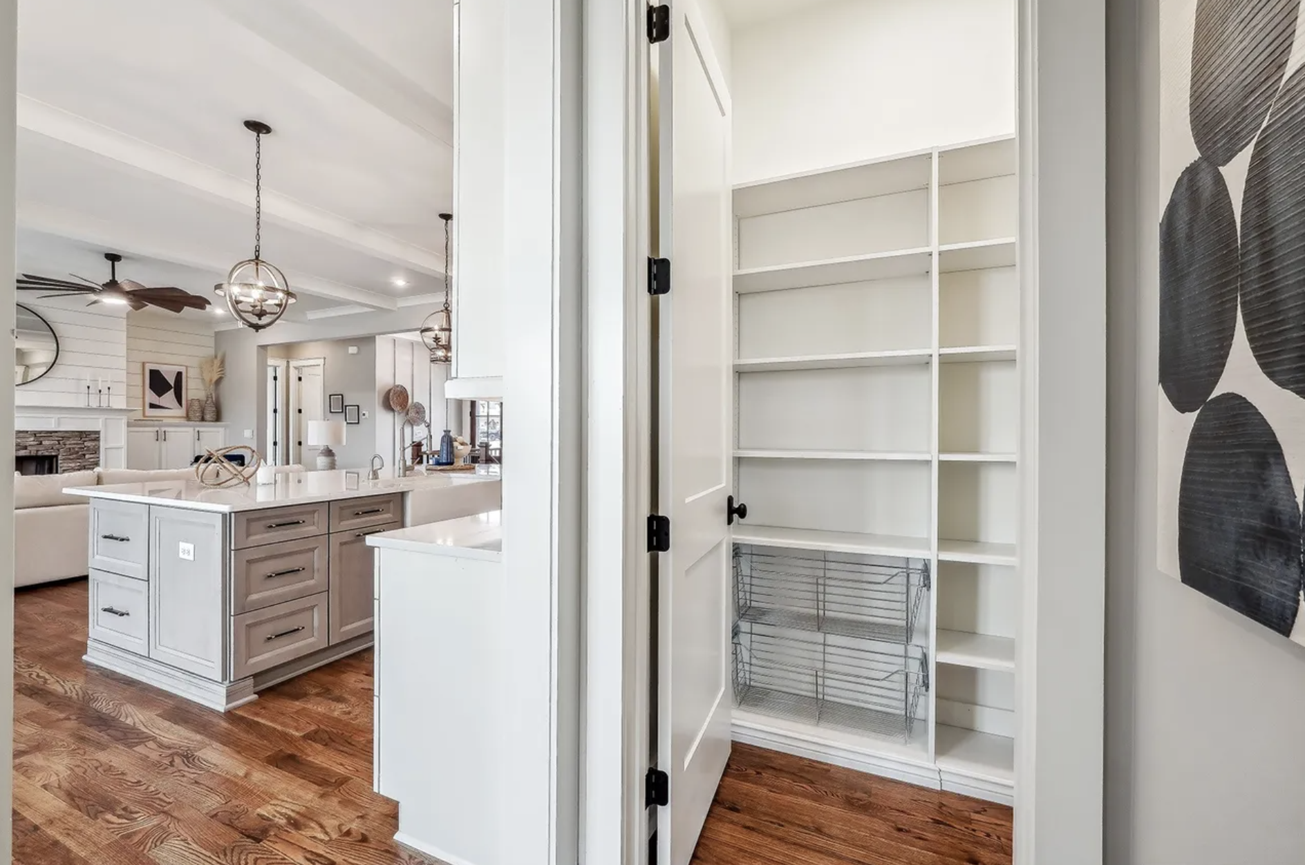 Empty pantry with white shelves and wood floor, adjacent to a kitchen with white cabinets and brown flooring, in a modern home.