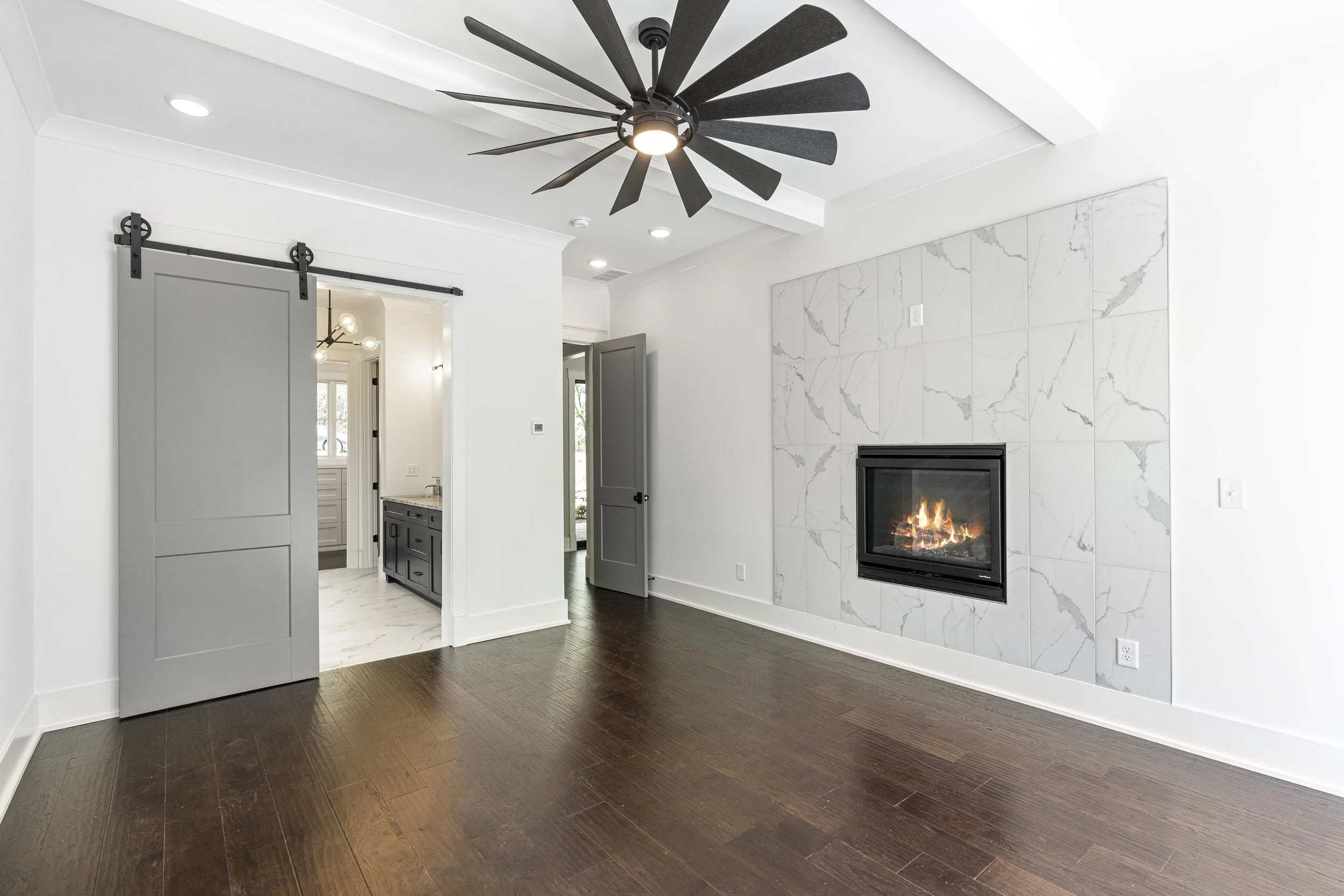 Living room with white walls, a modern fireplace with a marble surround, dark hardwood floors, a black ceiling fan, and gray sliding barn doors.