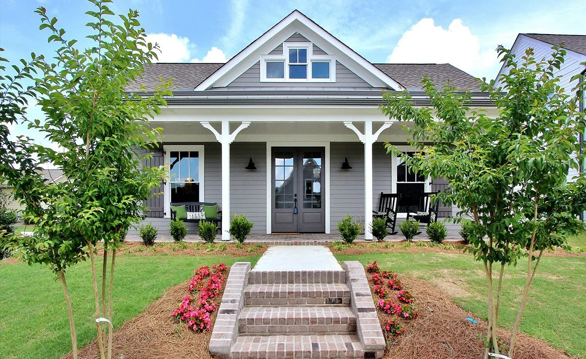 Front porch of a modern gray house with a gabled roof, white trim, and a single door. Brick steps lead to the porch, which has two black rocking chairs and a small sitting area with green cushions. There are trees and flower beds with pink flowers in