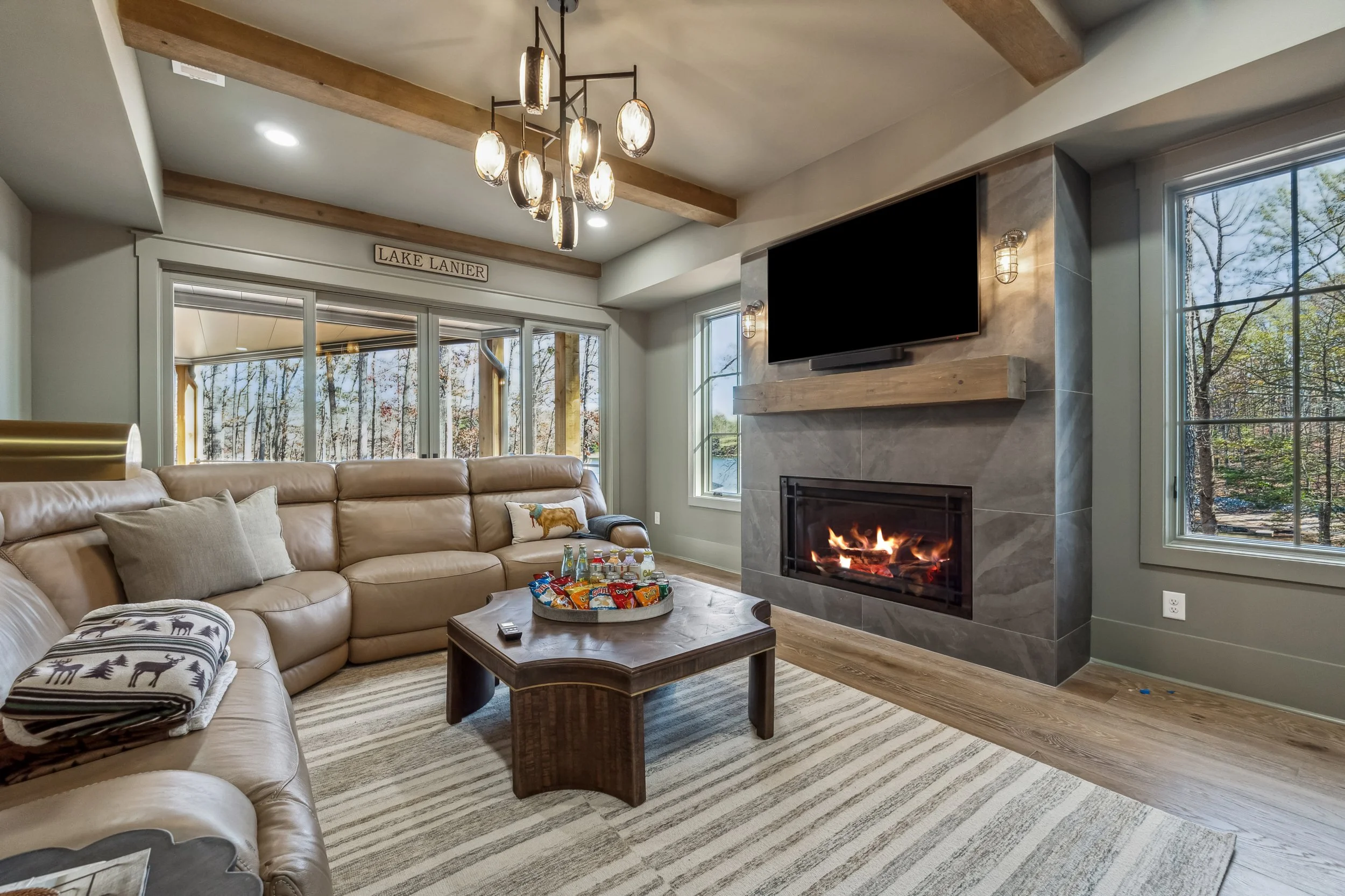 Living room with beige leather sofa, wooden coffee table with snacks, fireplace, large windows showing trees, flat-screen TV above fireplace, modern chandelier, and a striped area rug.