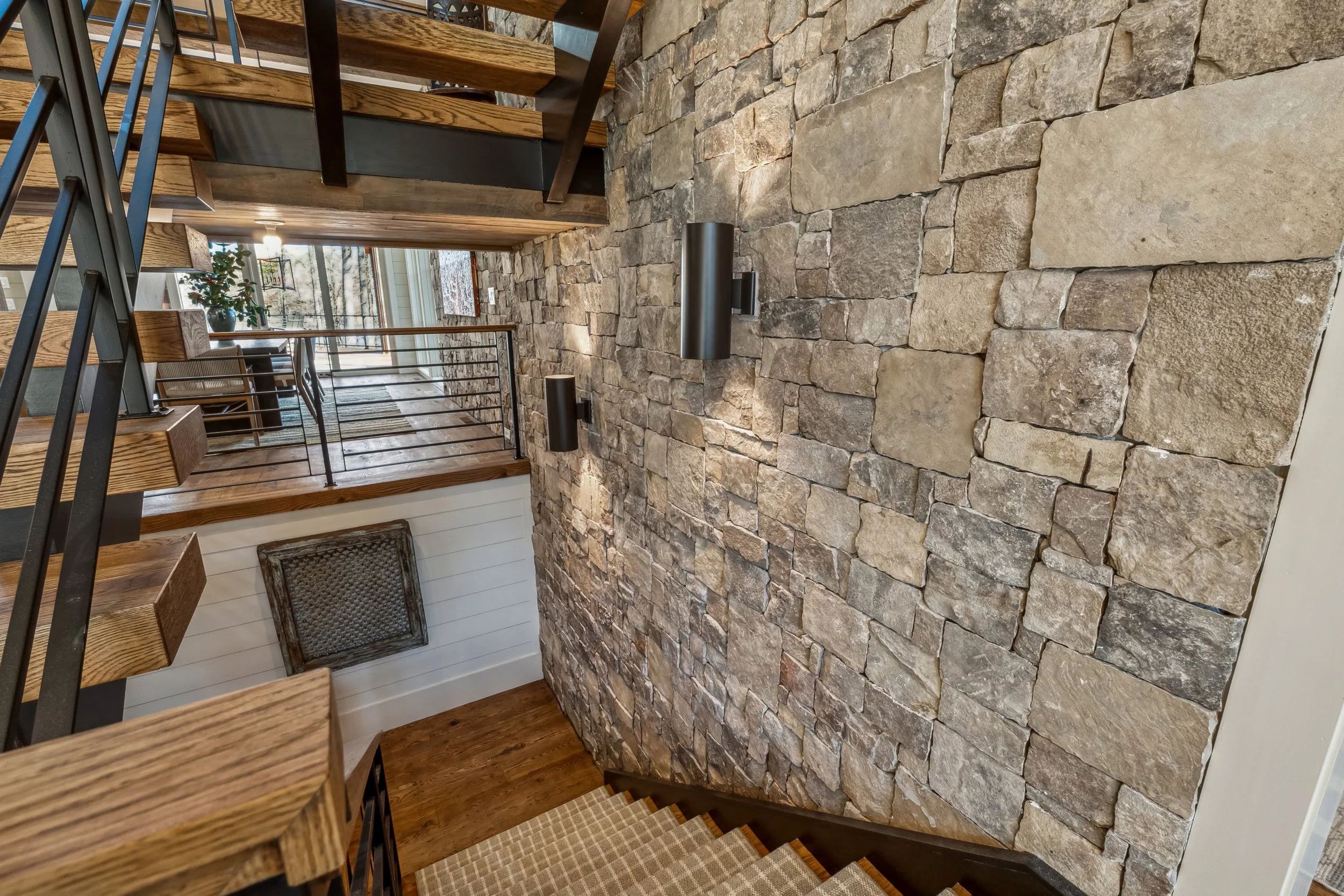 View of a staircase with wooden steps and black metal railing, a stone accent wall, and a glimpse of a dining area with a plant and large windows with a view of the outdoors.