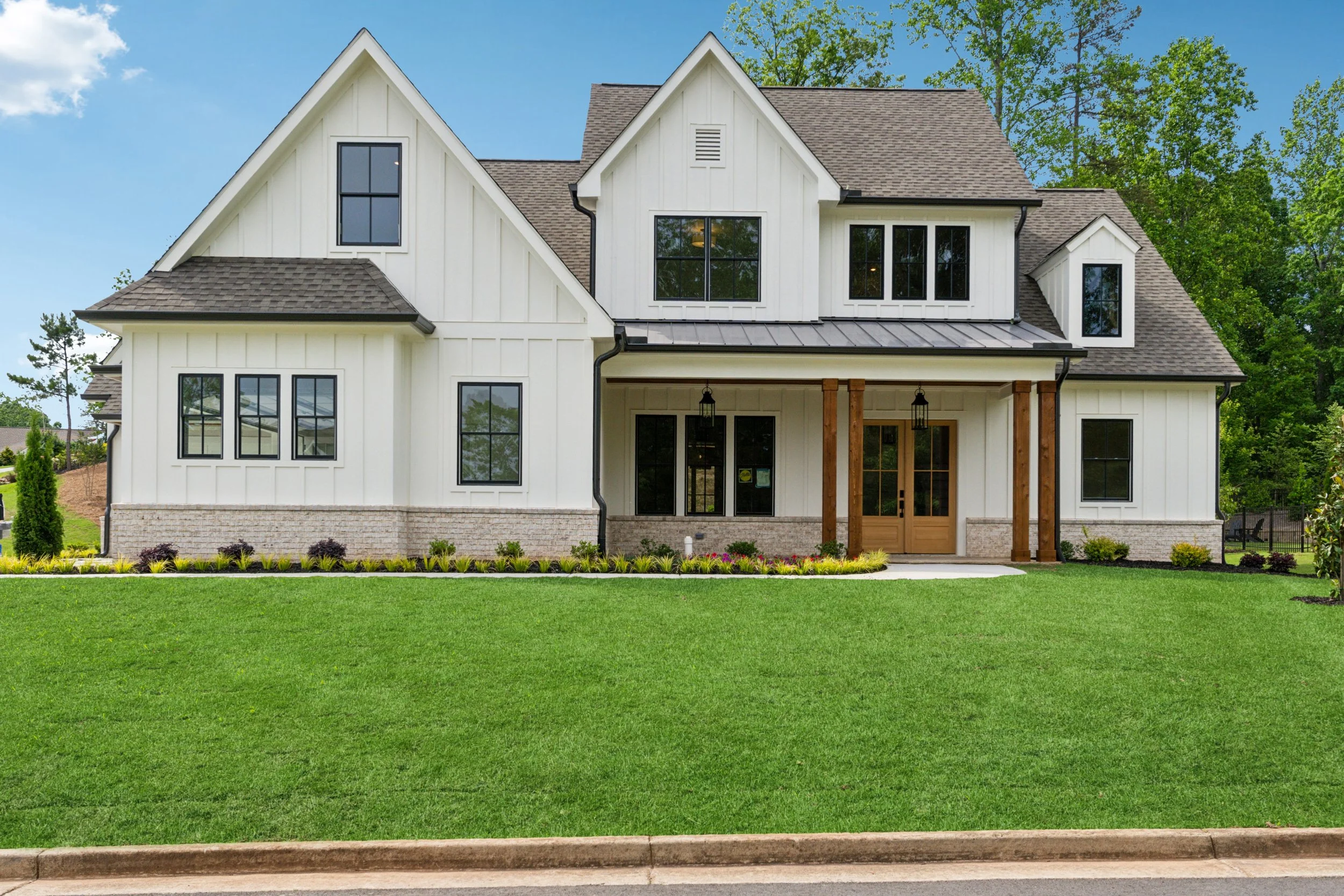 A modern two-story house with white exterior and black window frames, featuring a covered porch with wooden columns, surrounded by a green lawn, trees, and a clear blue sky.