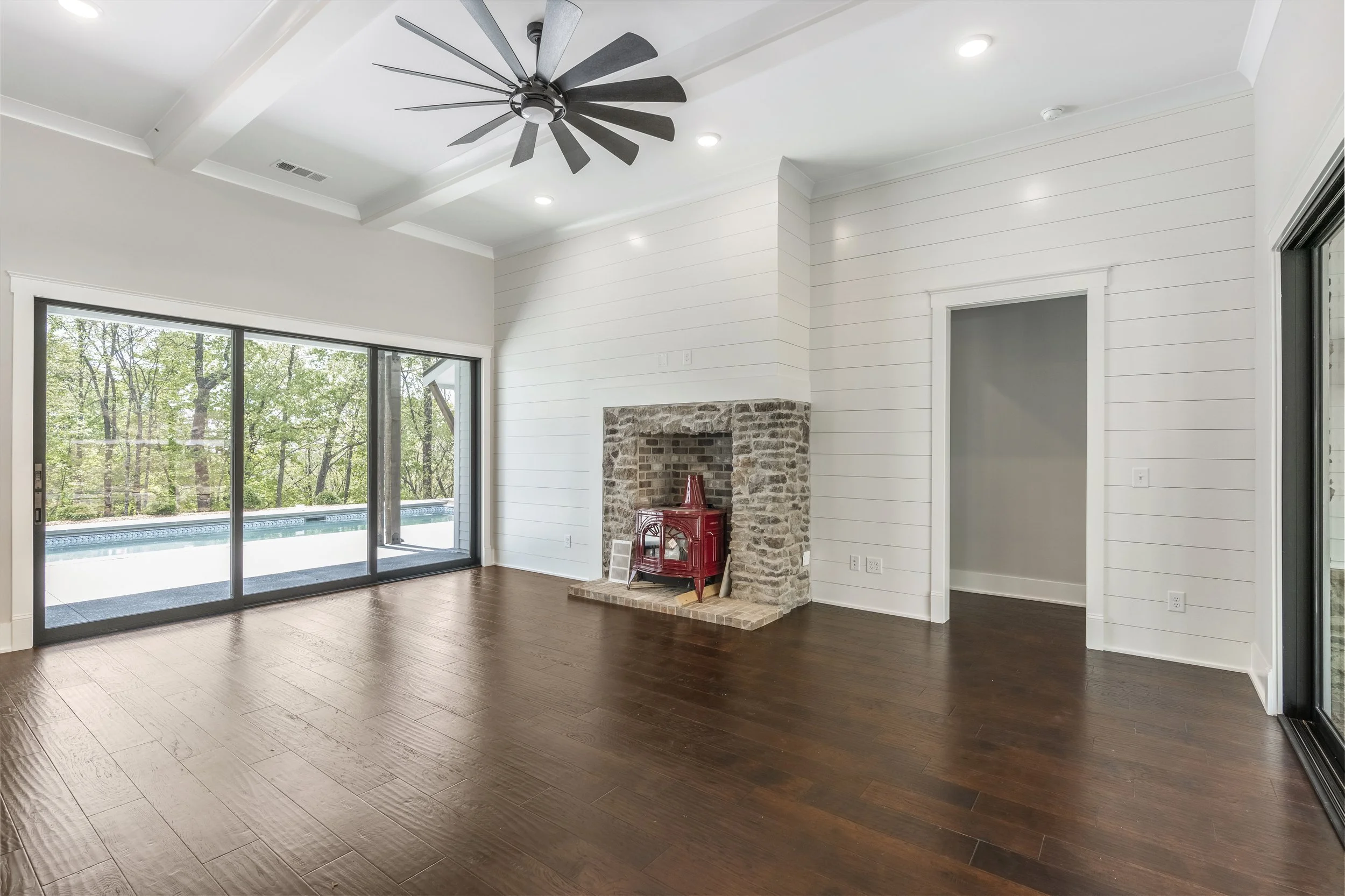 Living room with large sliding glass doors leading to a backyard with a pool, a brick fireplace with a small red stove, crown molding, recessed lighting, and wood flooring.