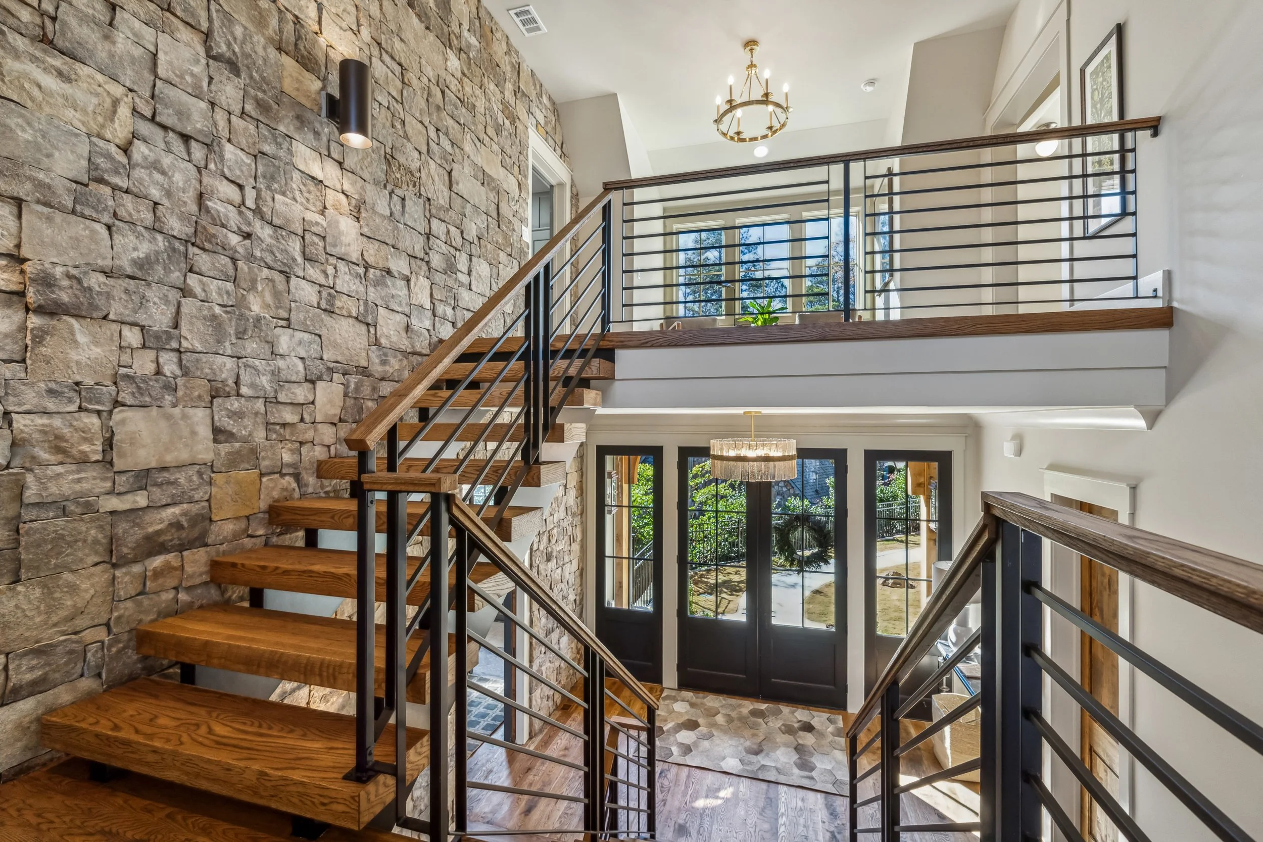 Interior view of a modern staircase with wooden steps, metal railings, a stone accent wall, and large windows providing natural light.